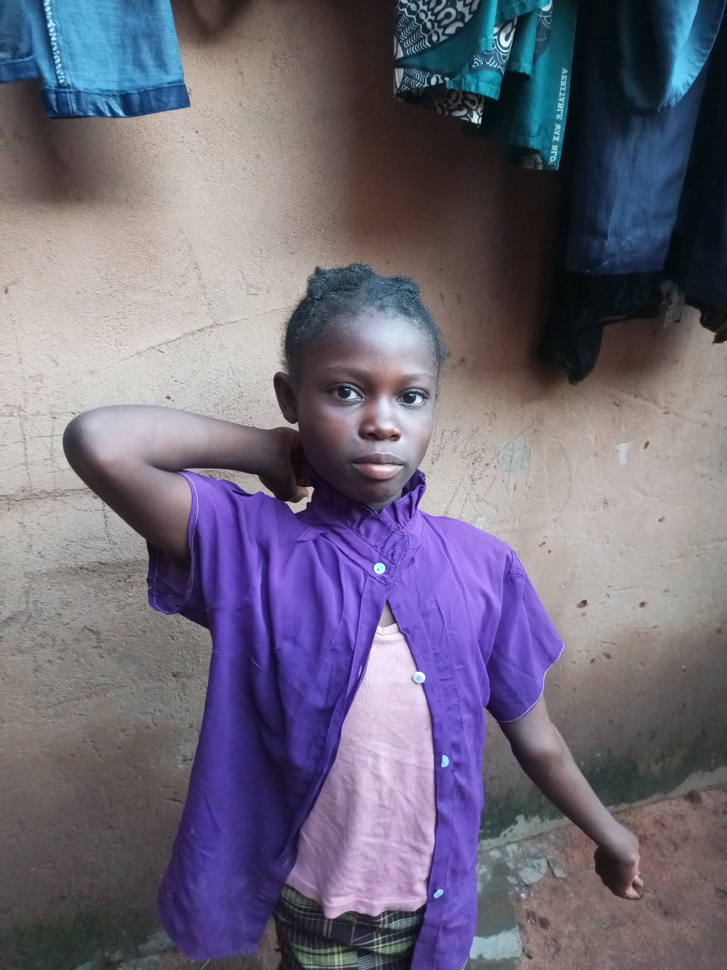 A young girl, with dark skin and dark braided hair pulled back, stands in front of a textured, light brown wall in Gombe, Nigeria. She is wearing a purple, short-sleeved, open-front shirt over a light pink undershirt. The purple shirt has a ruffled collar and is fastened with white buttons. Below her purple shirt, she appears to be wearing plaid shorts or a skirt in green and brown. Her right arm is bent, with her hand resting on the back of her head, and her left arm hangs down by her side, her hand in a fist. Clothing hangs from a line above and to the left of the girl, including what appears to be a pair of denim shorts and a patterned green fabric. The ground appears to be made of dirt and concrete.