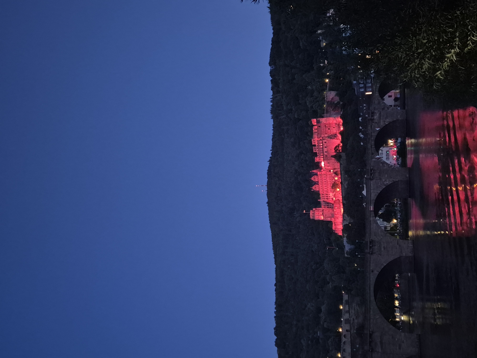This is a nighttime photograph of Heidelberg Castle illuminated in red, situated on a forested hill overlooking a river. A stone bridge with multiple arches spans the river in the foreground. The castle's red lighting casts a reflection on the water. The sky is a deep, clear blue. The scene captures the illuminated castle, the bridge, the river, and the surrounding hills, creating a picturesque and somewhat dramatic evening view of the city.