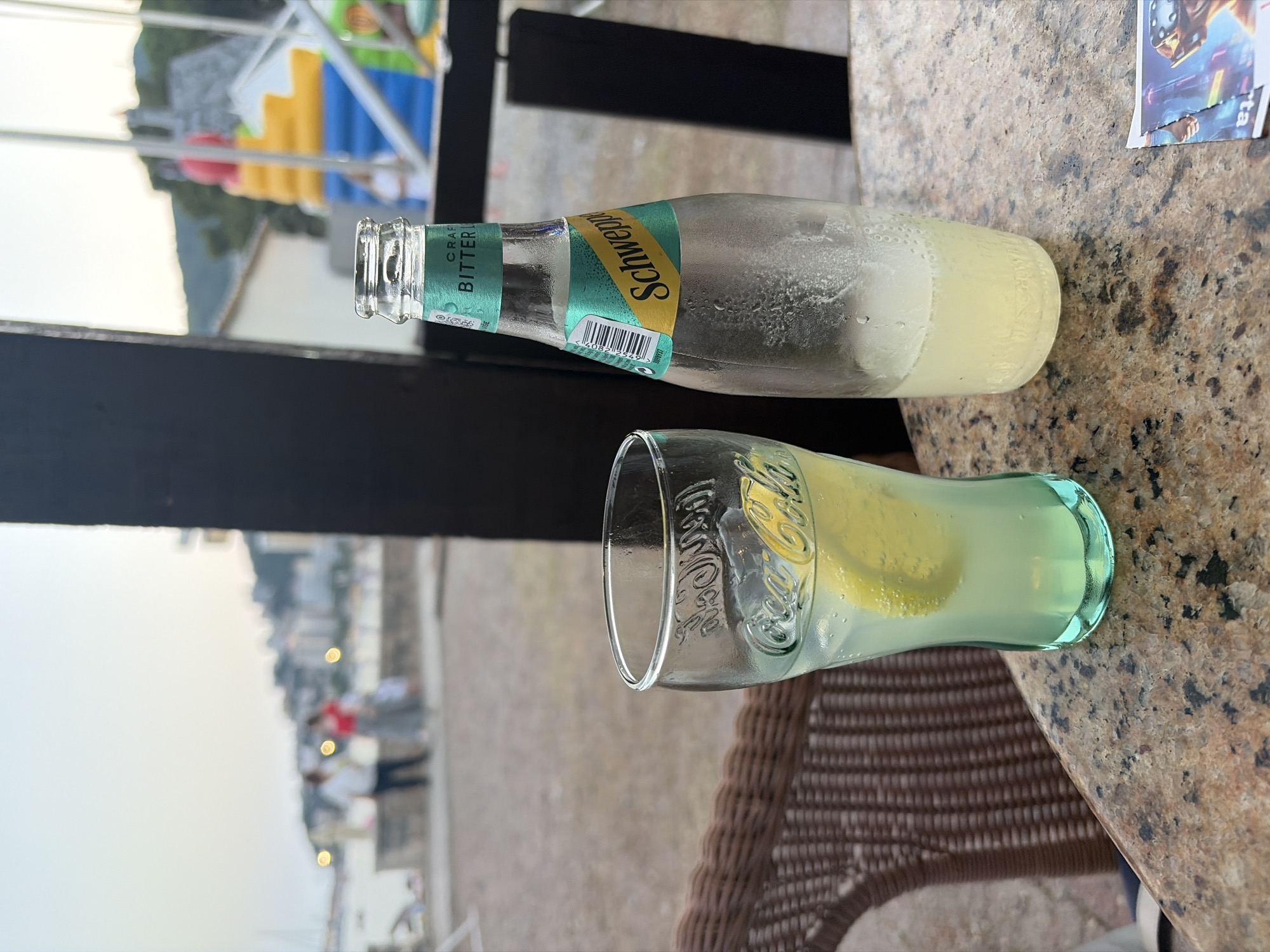 A still life photograph features two beverages on a speckled granite tabletop. To the left, a partially filled glass bottle of Schweppes Bitter Lemon lies horizontally. The bottle has condensation on its exterior and a prominent yellow label with the Schweppes logo. Visible text on the label includes "CRAFT," "BITTER," and a barcode. To the right, a clear glass with a pale yellow liquid and lemon slices rests vertically. The glass has embossed lettering that appears to read "Coca-Cola" around its rim.
In the background, out of focus, is a blurred scene of buildings and people, suggesting an outdoor setting, possibly a street or promenade. The lighting indicates it is daytime, with a soft, diffused light suggesting either early morning or late afternoon. The overall mood is casual and relaxed, with the focus on the refreshing drinks. The location context points to Budva, Montenegro, which could imply a coastal or urban environment.