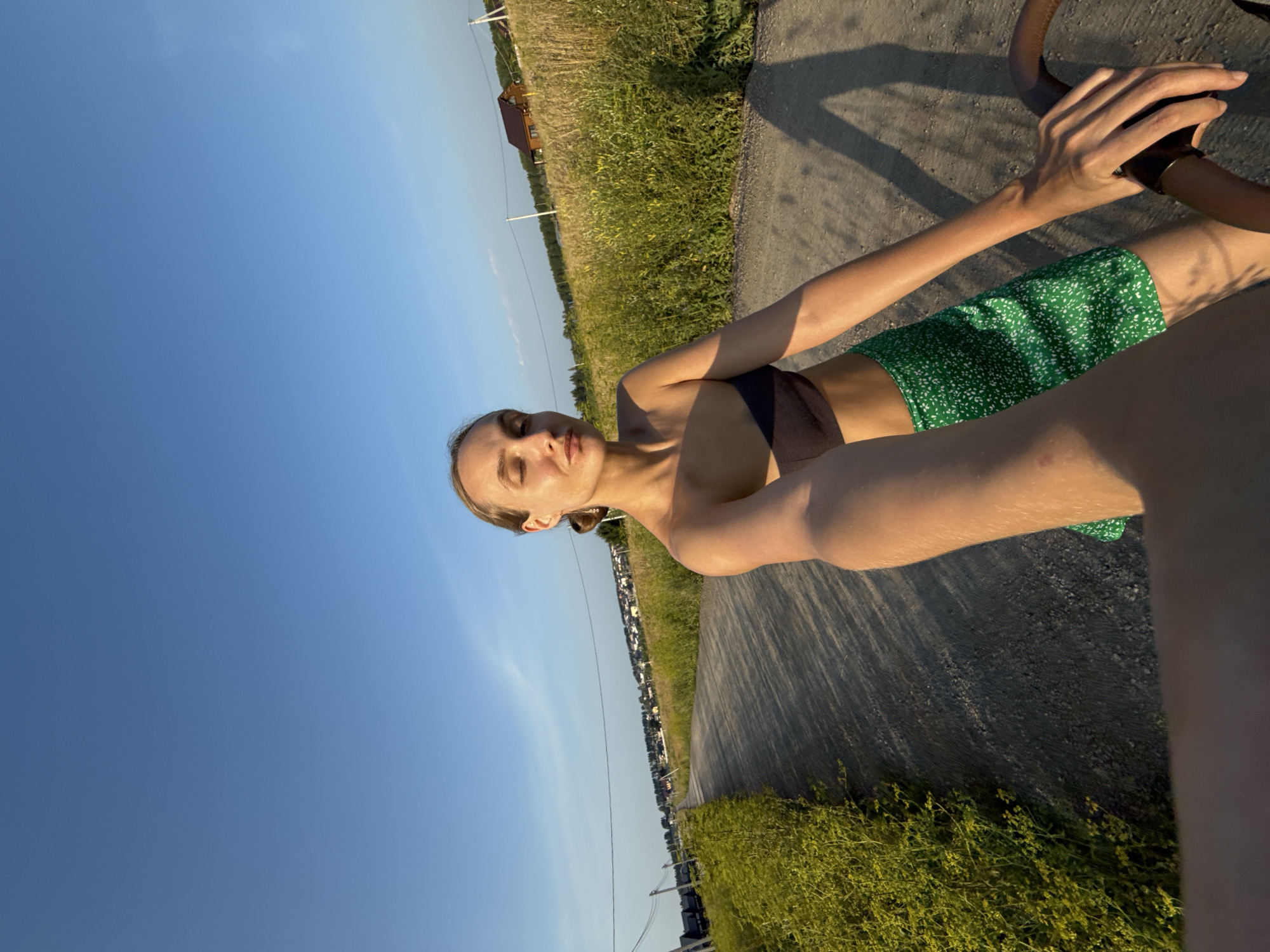 A young woman is captured from the chest up, riding a bicycle. She is wearing a dark sports bra and green shorts with a small white floral pattern. Her eyes are closed, and her head is tilted slightly upwards, suggesting a relaxed or perhaps meditative state. She is holding onto the handlebars of the bicycle with her right hand.
The scene is set on a rural road, with tall, green grass and wildflowers lining the right side. In the background, a small, wooden house with a dark roof is visible amidst the greenery. Beyond that, a line of trees and houses suggests a small village or settlement. The sky is a clear, vibrant blue, indicating a sunny day. The lighting suggests it is late afternoon or early evening, with long shadows cast on the road.
The image appears to be a selfie taken from the perspective of someone riding the bicycle. The overall mood is peaceful and serene, capturing a moment of enjoyment in a natural, rural setting in Podshivalovo, Russia. No visible text is present in the image.