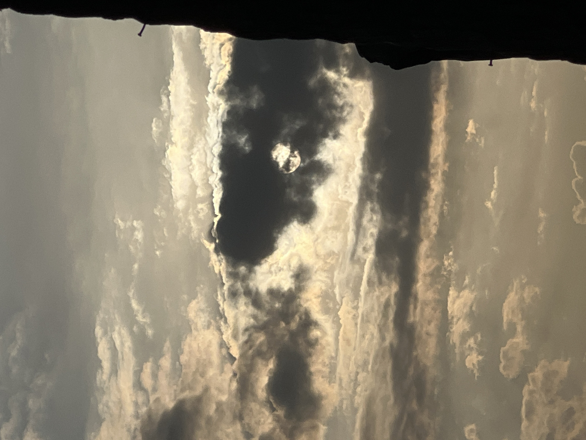 The image captures a cloudy sky with the sun partially obscured by dark clouds, creating a dramatic lighting effect. A dark silhouette of a building or structure dominates the right side of the frame, with a small, nail-like object protruding from its surface. The sky is filled with various cloud formations, some appearing bright and illuminated by the sun, while others are dark and dense. The overall tone is subdued and atmospheric. This scene is not specified to be in Kuta, Indonesia.