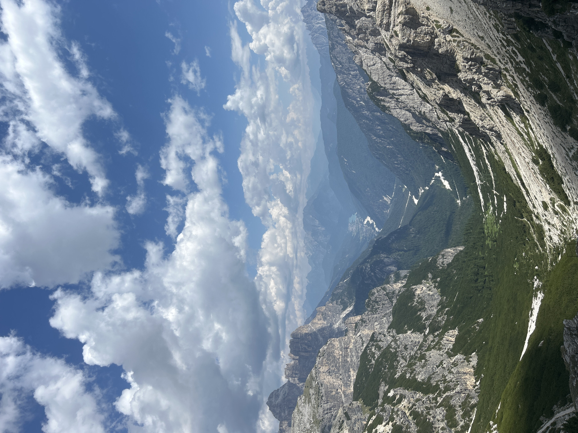 This is a landscape photograph taken from a high vantage point, looking down into a deep valley. The valley floor is covered with dark green trees, and a winding river or road is visible in the distance, shimmering white. The valley walls are steep and rugged, with layers of rock and patches of vegetation. In the foreground, the terrain is rocky and sparse, with some patches of snow. The sky is a vibrant blue, dotted with fluffy white clouds. The overall impression is one of vastness and natural beauty, with the rugged mountains dominating the scene. The scene appears to be in a mountainous region.