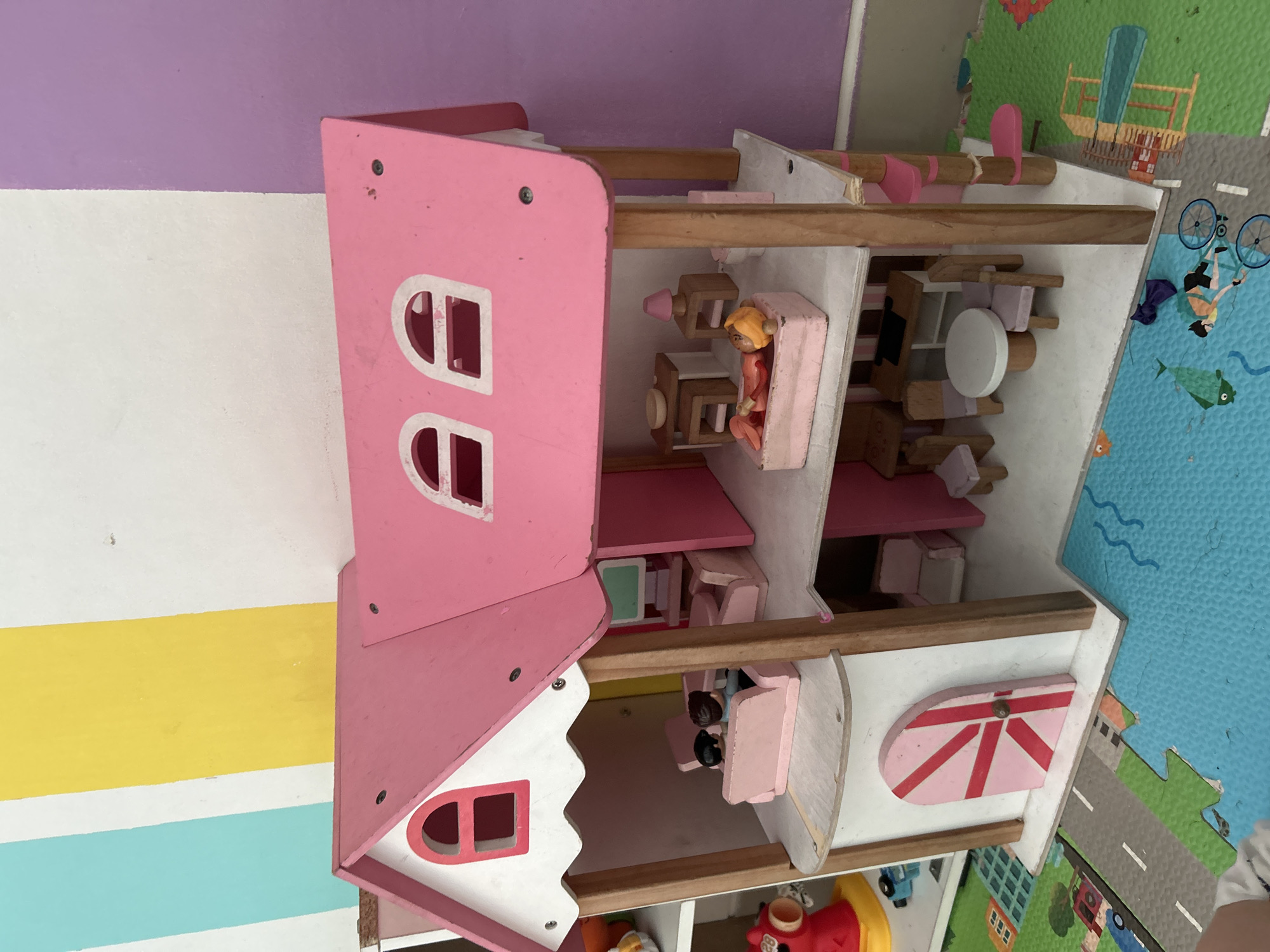 The image shows a pink dollhouse with multiple levels and rooms, situated on a colorful play mat depicting roads and water. The dollhouse features a pink roof with arched windows and a white facade. Inside, miniature furniture and figurines are visible, including a living room scene with a pink sofa and two figures, and a bedroom with a pink bathtub and a figure lying in it. There is also a dining area with a table and chairs, and a desk. The background consists of a wall with vertical stripes of light blue, yellow, white, and purple. The scene appears to be in a child's play area.
