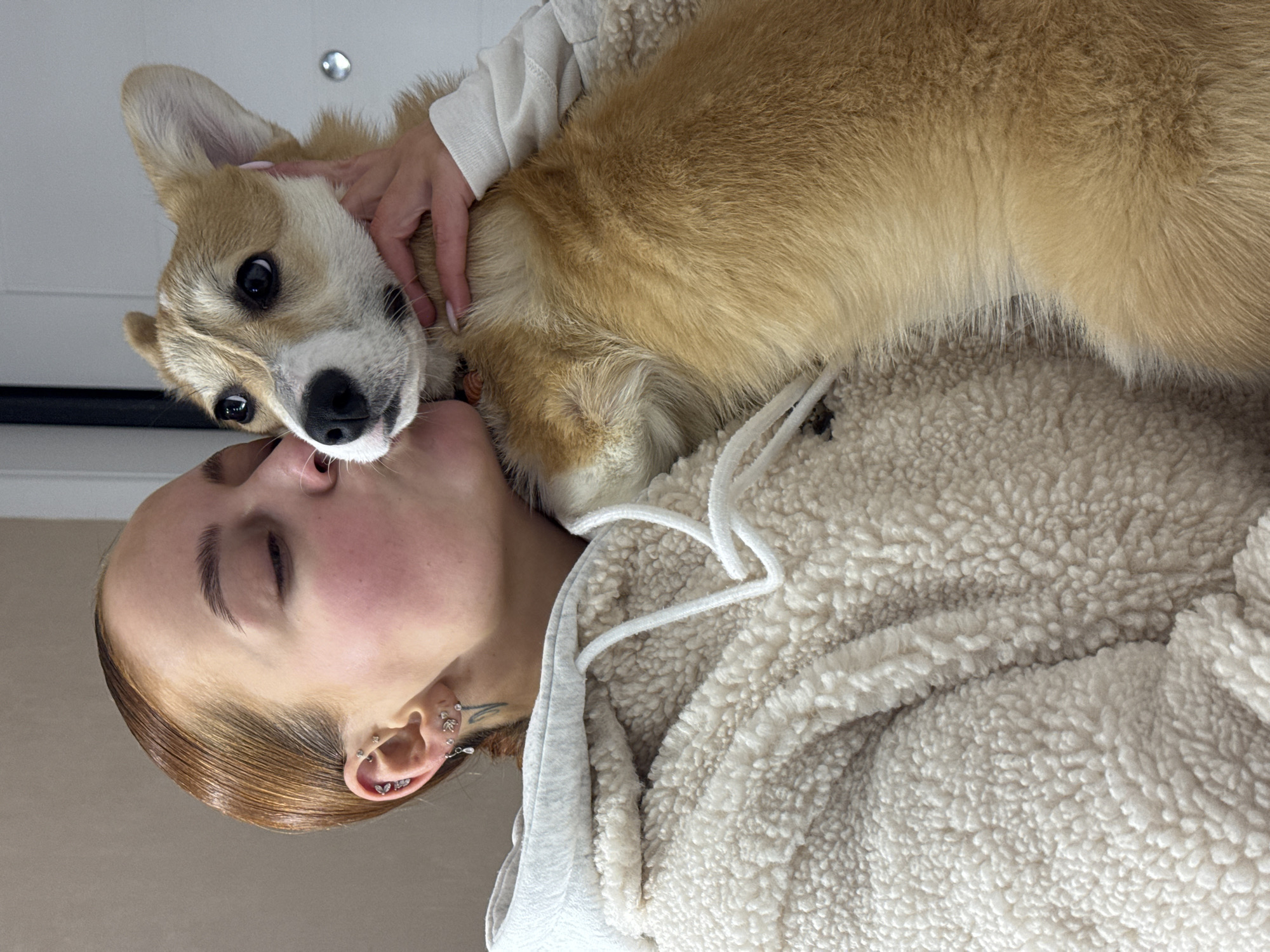A light-skinned woman with short, light brown hair and multiple earrings is seen tilted horizontally, cuddling with a tan and white corgi dog. The woman is wearing a cream-colored, sherpa-textured hoodie with white drawstrings. The dog rests on the woman's chest. The location is not identifiable.
