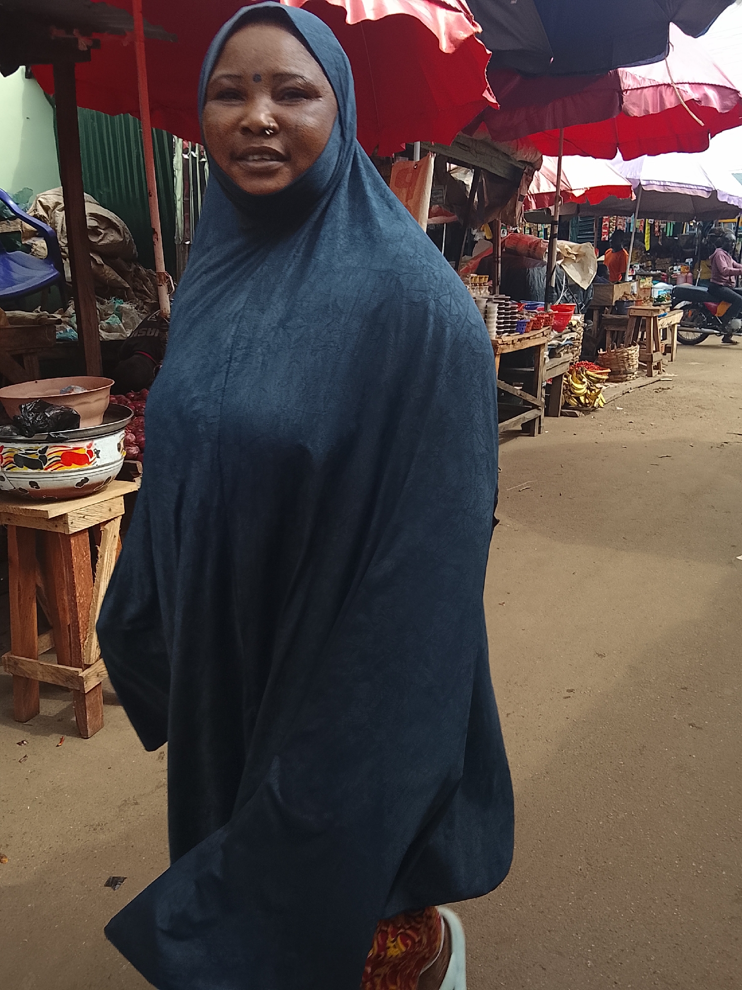 In the city of Akwanga, Nigeria, a woman wearing a dark blue hijab stands in a marketplace. She has a bindi on her forehead and a nose ring. She is looking towards the camera with a slight smile. The background shows stalls with various goods, including produce like bananas and red onions. Colorful umbrellas provide shade over the stalls. In the distance, a person on a motorcycle is visible, and other market-goers can be seen. The ground is a dirt path, typical of an outdoor market. The overall scene captures a moment of daily life in a Nigerian market.