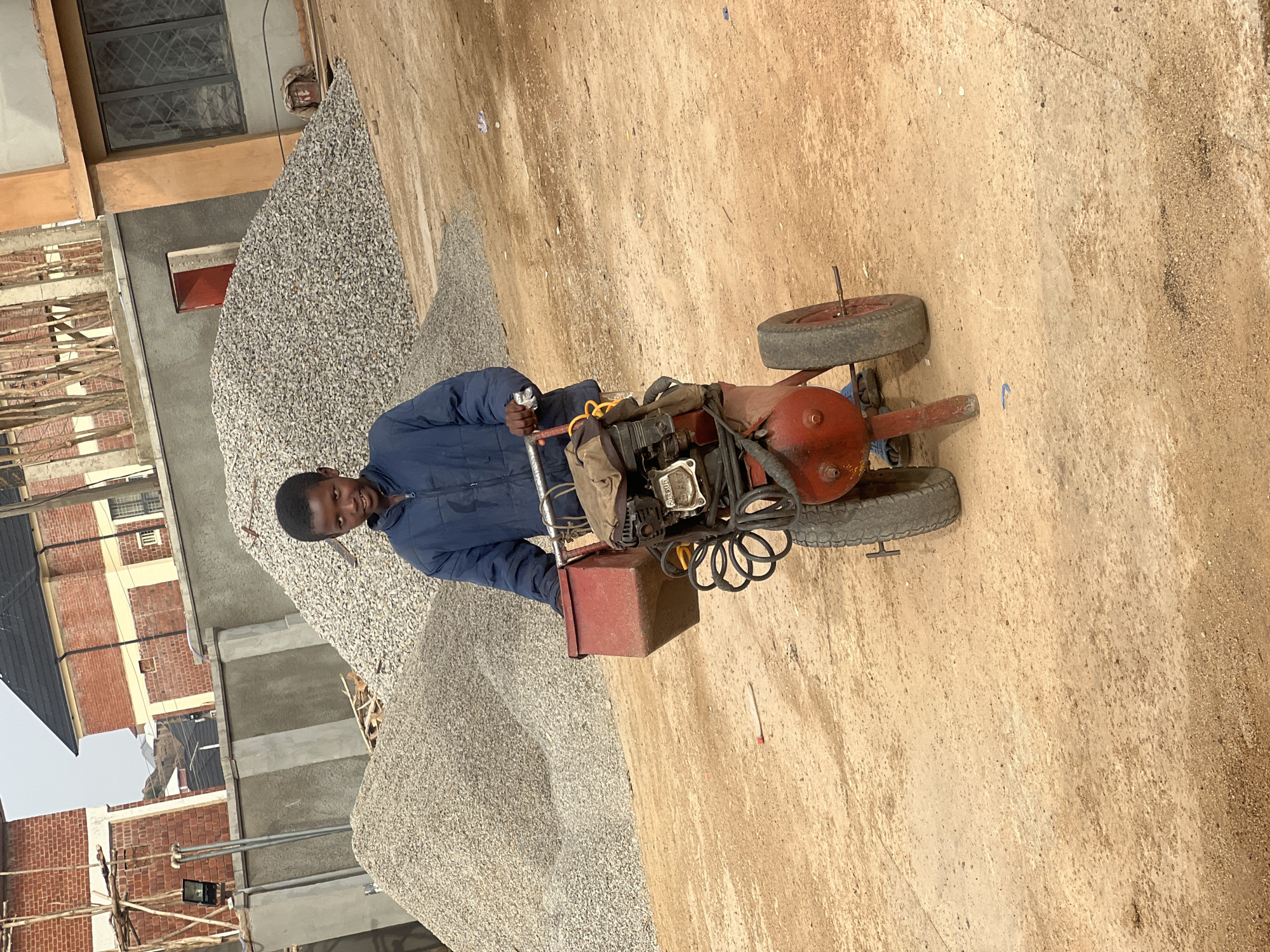A young male stands in a dirt area in Barakin Lerawi, Nigeria, next to a pile of gravel. The male is wearing a navy-blue jacket. He is standing behind a small, red motorized machine with four wheels. A red metal box is attached to the side of the machine. The gravel pile sits next to a building under construction.