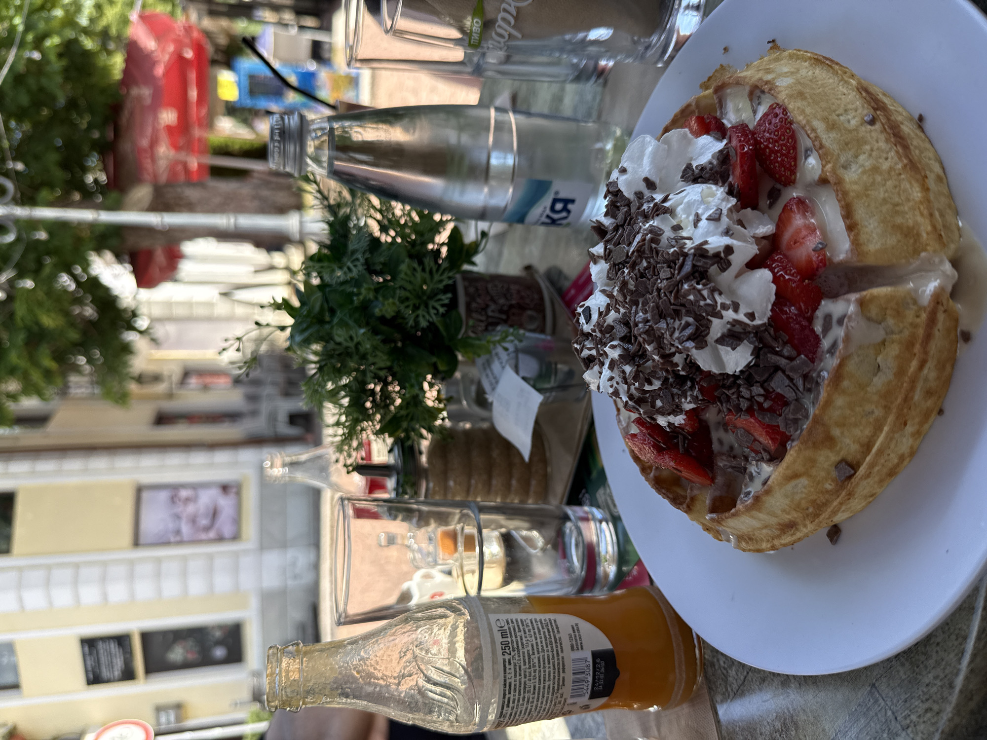 The image shows a dessert plate on a table, likely at an outdoor café. The dessert appears to be a waffle topped with whipped cream, sliced strawberries, and chocolate shavings. Beside the plate are a glass of water, a bottle of mineral water labeled with Cyrillic script, a small potted plant, a glass, and a bottle of orange juice. The background shows a street with buildings, indicating an urban setting. Based on the "Lovech, Bulgaria" prompt, it can be inferred that the café is located in Lovech, Bulgaria. The scene suggests a pleasant outdoor dining experience in a European city.