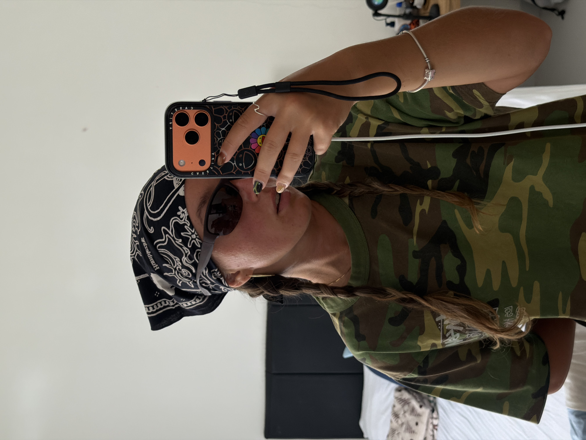 A person takes a selfie in an indoor setting. The individual has braided brown hair, a black bandana on their head, and sunglasses. They are wearing a camouflage shirt. The phone in their hand is in a black case with a flower design. The location is unknown, but the prompt suggests it could be in Kuta Selatan, Indonesia. The background includes a white wall and a partial view of furniture.