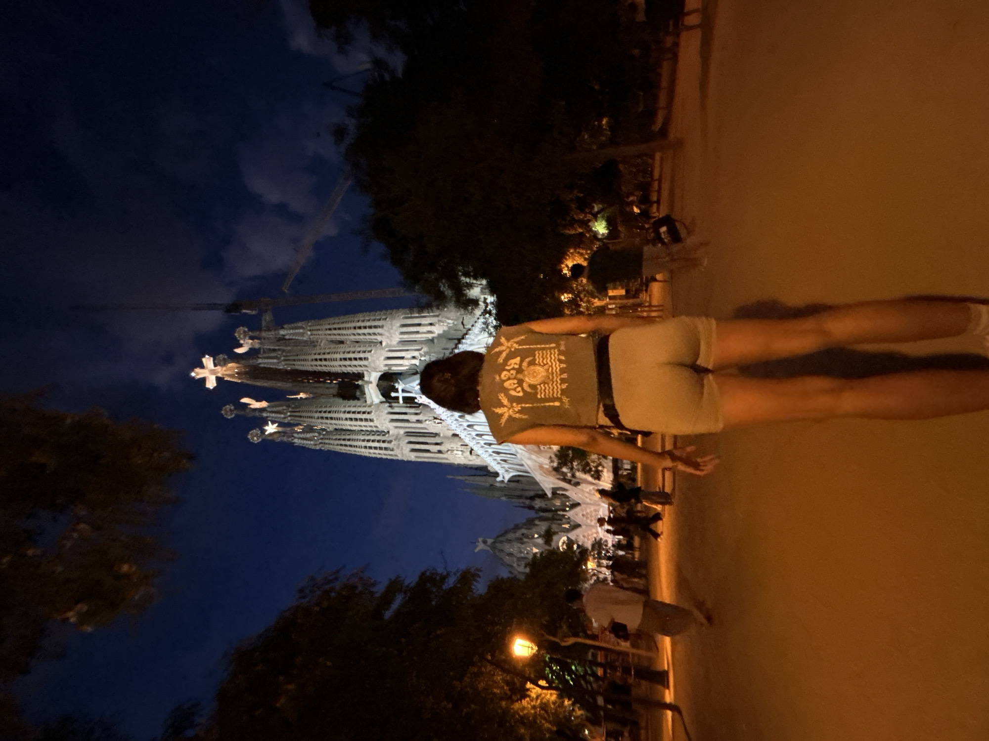 This is a nighttime, outdoor, full-body shot of a woman from behind, walking away from the camera towards the Sagrada Familia basilica in Barcelona, Spain. The basilica is illuminated and prominent in the background, with cranes visible behind its spires, indicating ongoing construction. The sky is dark blue with scattered clouds. The woman is wearing a sleeveless shirt with a graphic print and light-colored shorts. Her legs are visible from below the shorts. She has dark hair. The ground in front of her is a sandy or dirt path illuminated by orange streetlights. Other people are visible in the midground, walking in various directions, some near a lamppost on the left. Trees and foliage frame the scene on both sides.
