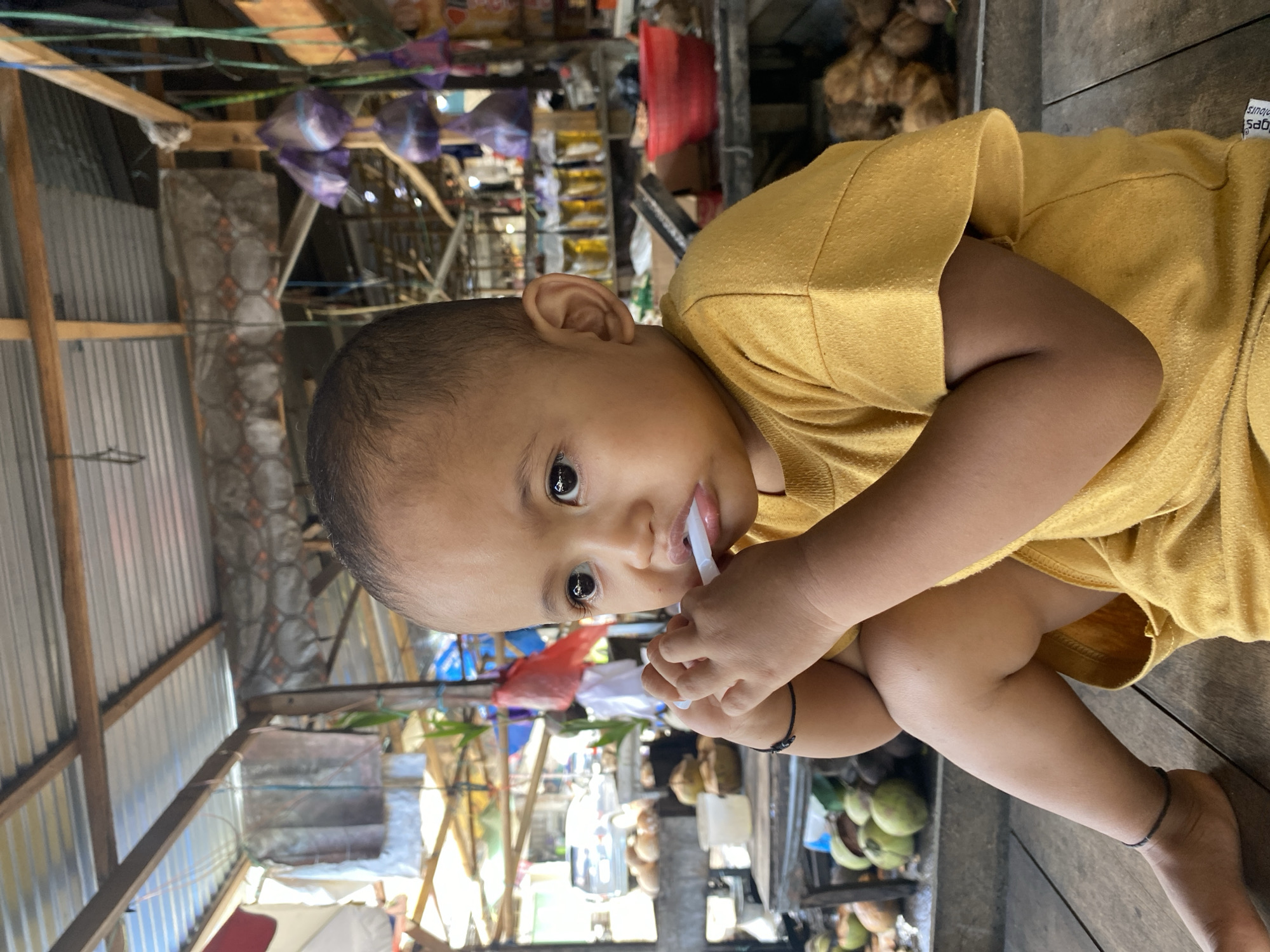 A young child, likely a toddler, is seated on a wooden surface, looking to their left with a curious expression. The child is wearing a yellow t-shirt. Their hands are raised to their mouth, holding a white object, possibly a pacifier or toy, which is partially in their mouth. The background suggests an outdoor market setting. A corrugated metal roof is visible above, supported by wooden beams. Various items are displayed in the background, including what appear to be coconuts, bags of produce, and shelves stocked with goods. The overall impression is that of a casual, everyday scene in a bustling marketplace. The image was taken in the city of Ta', Indonesia.
