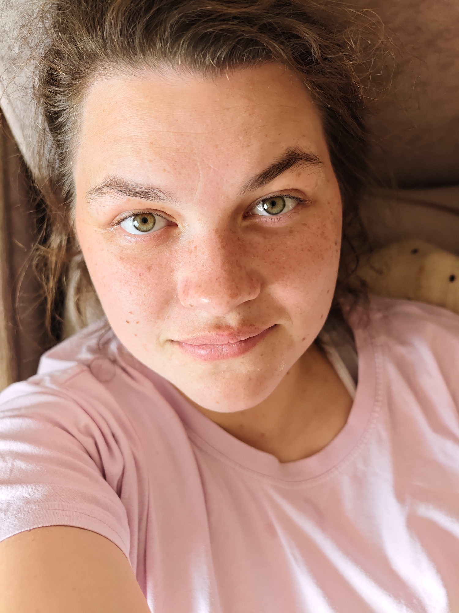 A close-up, high-angle selfie shows a young woman with fair skin and freckles. She has light brown, slightly curly hair pulled back from her face. Her eyes are green. She is wearing a light pink t-shirt. The background includes a beige surface. The location is potentially Уральское, Russia. No specific details confirm this location, it is speculative.