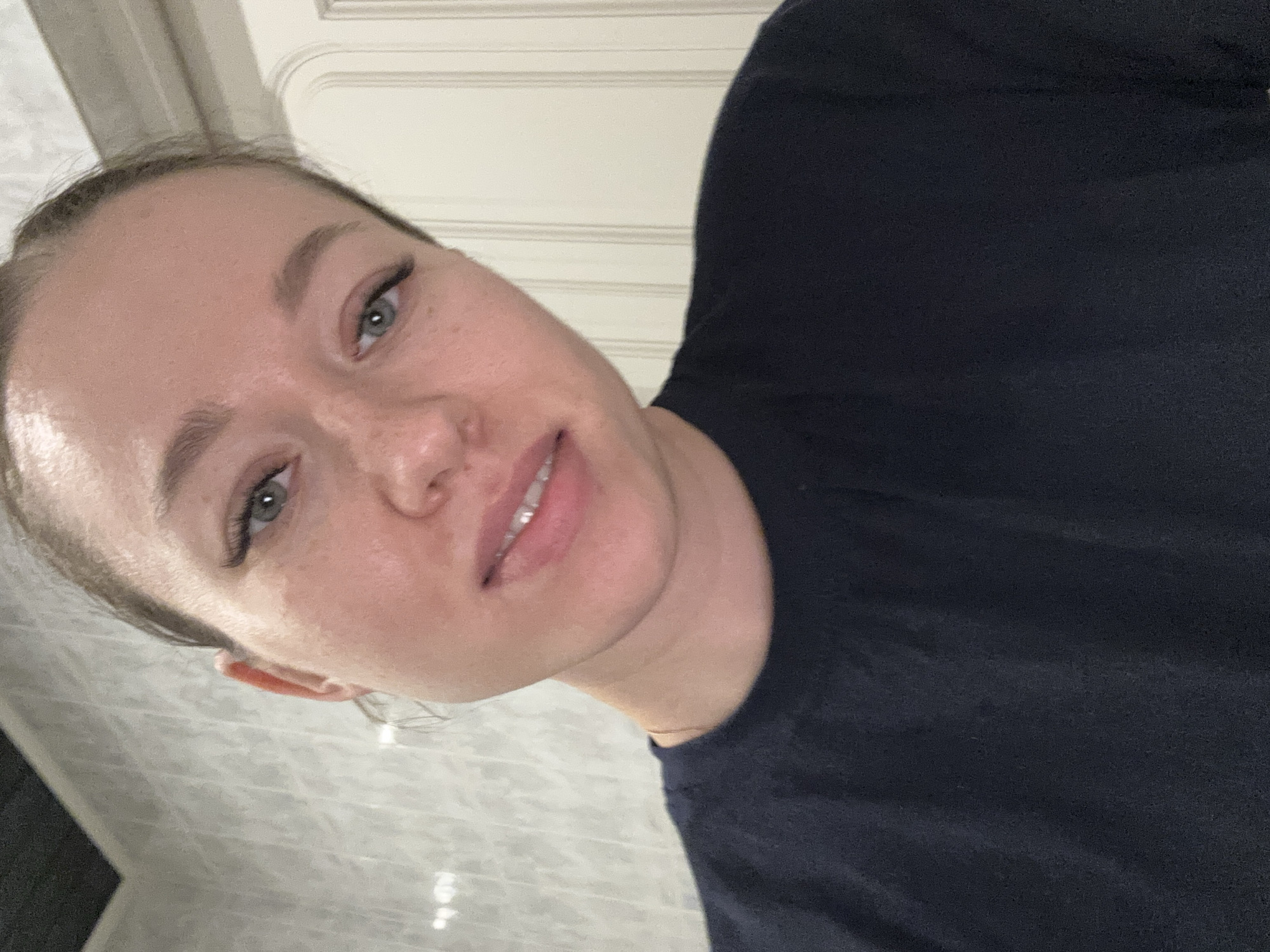 The image shows a fair-skinned woman with her face close to the camera. She has light gray-green eyes and freckles visible on her nose. Her hair is pulled back. She is wearing a dark blue shirt. The background consists of a door with horizontal panels. The location in the city of Ферма, Russia is not discernible.