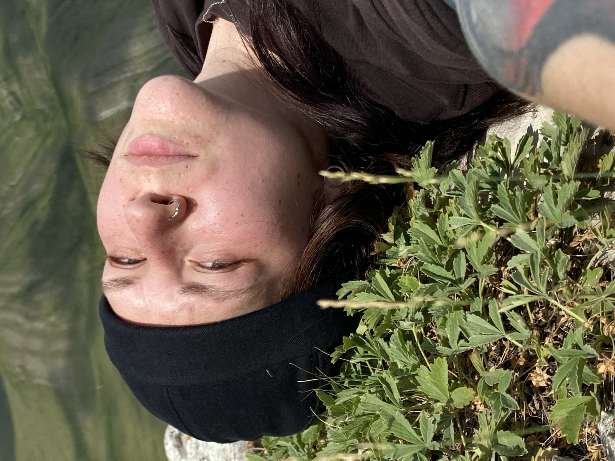 A person is lying down on the ground, looking up at the sky. They are wearing a black beanie and a brown shirt. Green plants are growing around them. In the background, there are rolling hills covered in green grass. The person has a nose ring. The location is the village of Абзаново, Russia.