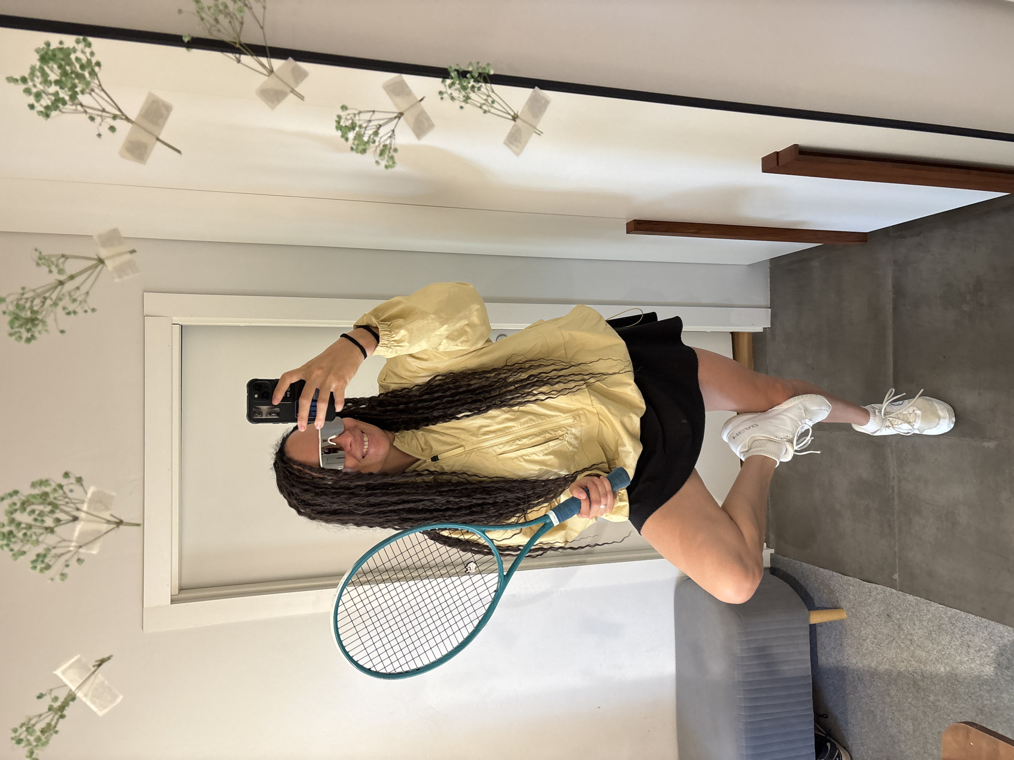 In a room with white walls, a person with long, dark dreadlocks is taking a selfie in a full-length mirror. They are wearing a yellow, hooded jacket, a black skirt, and white sneakers. The person is holding a teal tennis racket in their left hand and has their right hand up to take the photo with their phone. Their left leg is bent at the knee and resting on their right calf, and they are leaning slightly.
The room has a gray upholstered bench with wooden legs on the left side, and a gray carpet is laid out on the floor. The mirror on the right reflects the scene, including several sprigs of baby's breath taped to the wall. The overall impression is of a casual, possibly athletic setting.