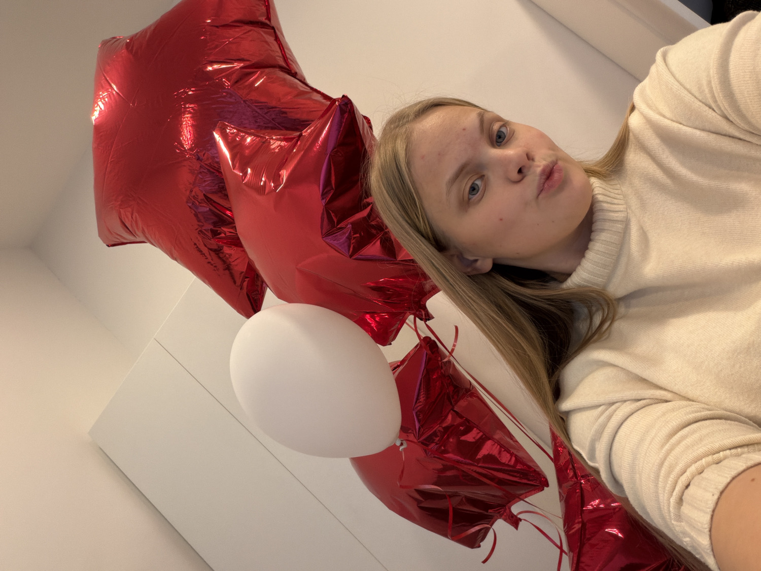 A young woman with long blonde hair, wearing a cream-colored sweater, takes a selfie with red star-shaped and a white round balloons. The background is a white wall, the photo appears to be taken indoors. There is no indication of Izhevsk, Russia.