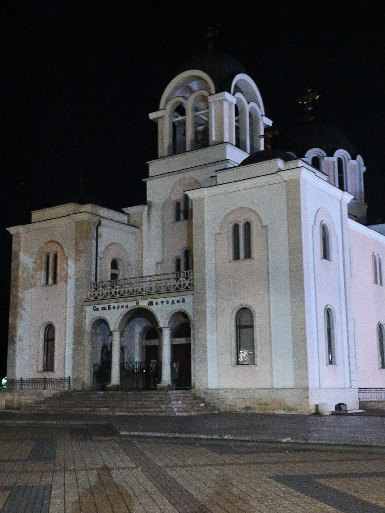 The image shows the exterior of the Church of Saints Cyril and Methodius in Lovech, Bulgaria, at night. The church is a large, multi-story building with a dominant central tower featuring arched windows and multiple domes, two of which are visible and topped with crosses. The facade is painted white with light pink accents and features arched windows and ornate decorative elements, including a railing with circular motifs above the entrance arches. A set of stone steps leads up to the main entrance, which is a portico with columns and arches. The surrounding area appears to be a paved public space with brick-like patterns, and the scene is illuminated by artificial light, casting shadows on the ground. The sky is dark and appears clear.