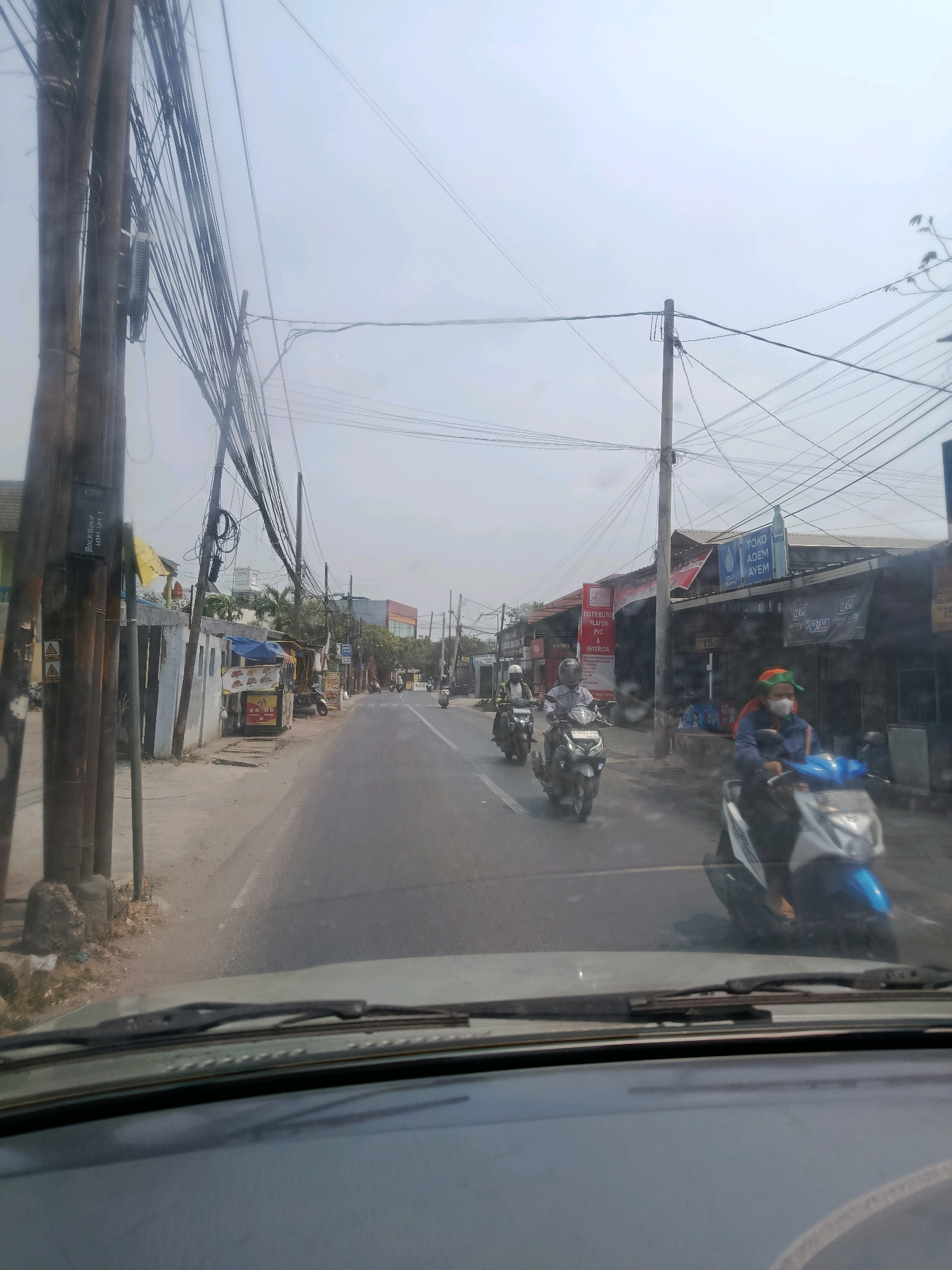 The image depicts a street scene in East Jakarta, Indonesia, characterized by a two-lane asphalt road. On the left side of the road, there are utility poles with a dense network of electrical wires, several small buildings, and a food stall. On the right side, there are more buildings, including shops with signage such as "TOKO ADEM AYEM" and other commercial establishments. Several motorcycles are visible on the road, with riders wearing helmets. One motorcyclist in the foreground is wearing a blue jacket and a green headscarf, and a face mask. The sky is overcast. The perspective is from inside a vehicle, with the windshield and dashboard visible in the foreground.