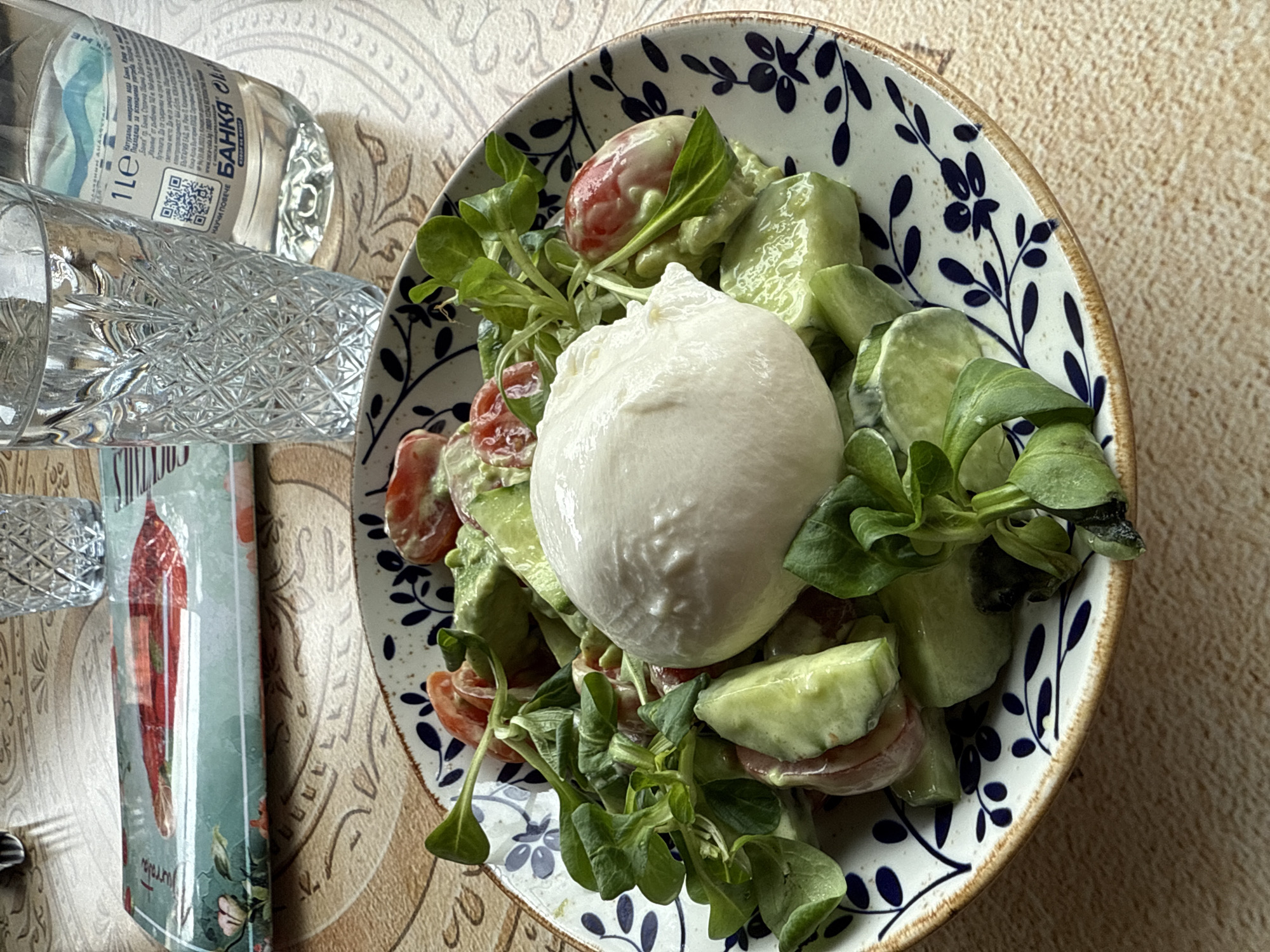 The image shows a partially visible table setting with a salad as the main subject. The salad, served in a white ceramic bowl with a blue floral pattern and a brown rim, appears to contain fresh greens, sliced cucumbers, halved cherry tomatoes, and a central ball of mozzarella cheese. A light dressing coats the salad components.
Adjacent to the bowl, to the left of the frame, are a partially full glass of water, a 1-liter water bottle labeled "Банкя" in Cyrillic script, and a cookbook with a stylized image of chili peppers on its cover. The presence of the “Банкя” bottled water strongly suggests the location is in Bulgaria, possibly in the city of Sofia, where Банкя mineral water is a common brand. The tabletop has a beige, patterned surface. No people are directly visible in the frame, focusing the scene on the food and surrounding items.