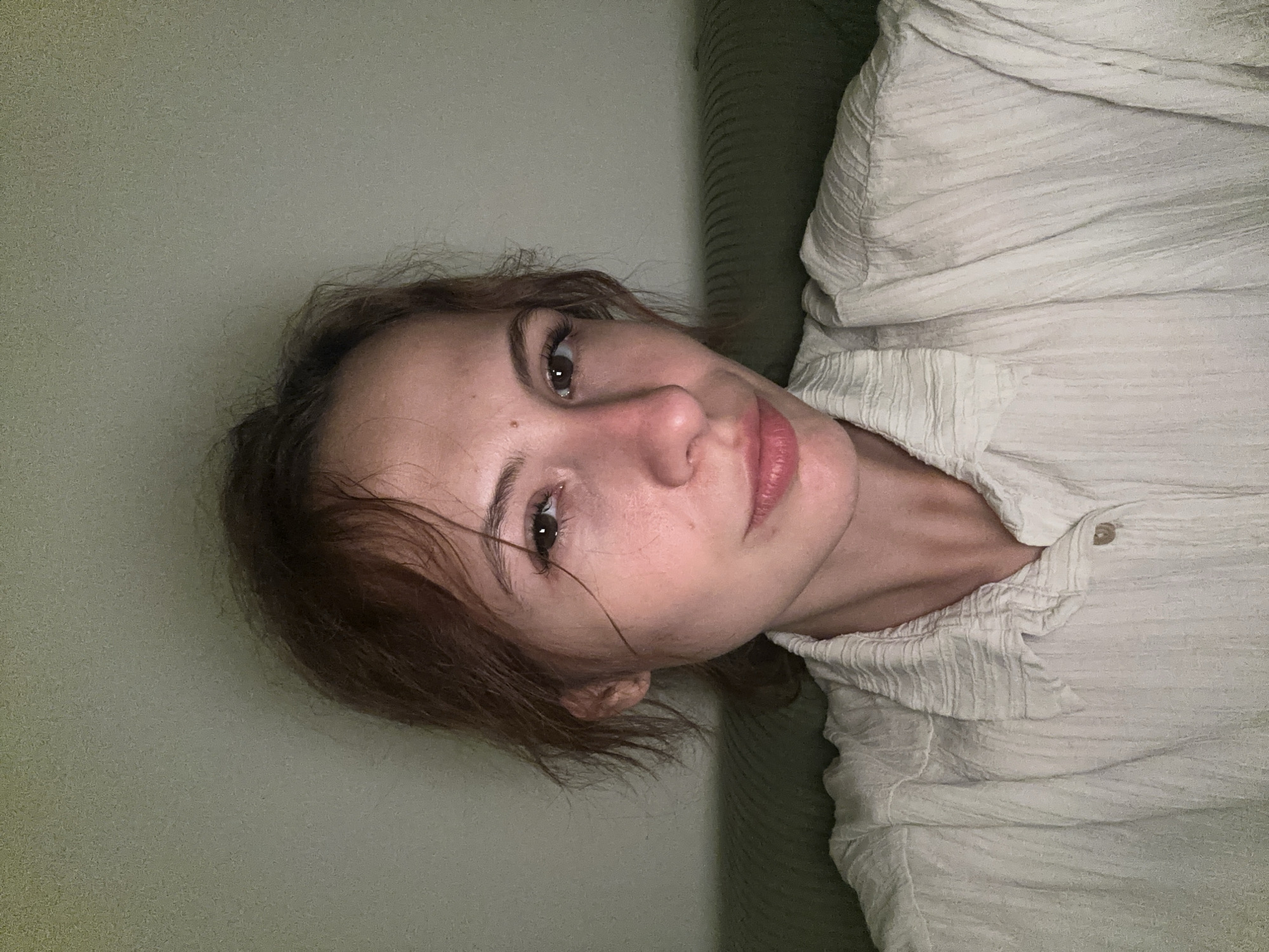 A person with brown hair is shown in a close-up, vertically oriented shot. The person is wearing a light-colored, textured shirt. Part of a light green wall or surface is visible on the left side of the image. The setting is likely indoors, but there are no specific location markers to indicate Novi Sad, Serbia. No specific actions or interactions are captured in the image.