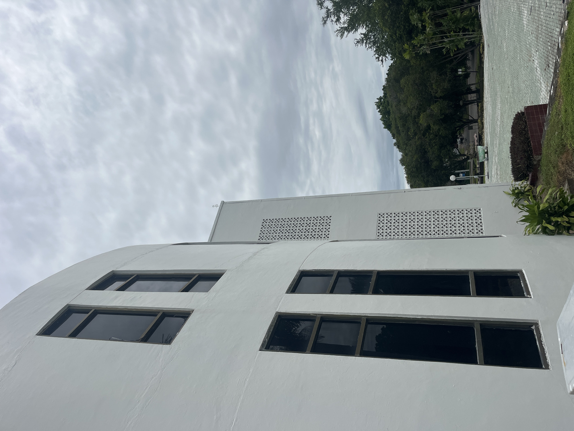 This is an exterior shot of the Islamic Center Lhokseumawe in Lhokseumawe, Indonesia. The image focuses on the lower portion of a white, multi-story building with a curved facade. Several rectangular windows with dark frames are visible. The building has sections with geometric latticework above the windows. In the background, a tree-lined area and a cloudy sky are visible. There are no people.