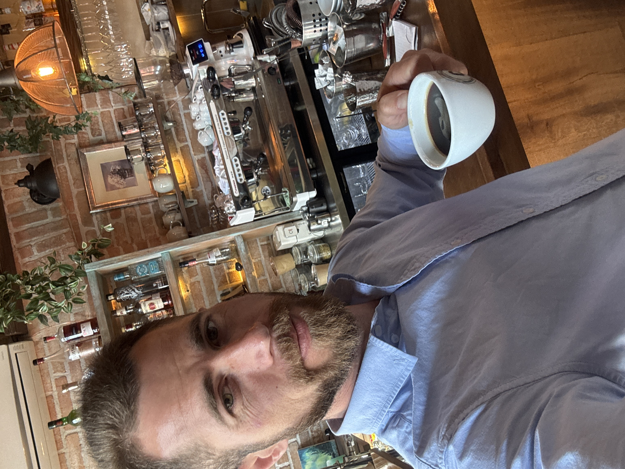 A man is captured in a close-up shot, holding a small white coffee cup filled with dark liquid, likely coffee, in his right hand. He has a short beard, short hair, and green eyes, looking directly at the camera with a neutral expression. He is wearing a light blue collared shirt.
The setting is an indoor commercial establishment, such as a cafe or bar, featuring a rustic brick wall as a prominent background element. To the man's left, a professional espresso machine, possibly a "Cremesso" model as suggested by partially visible text, is situated on a wooden countertop. The machine has multiple group heads and is surrounded by various coffee accessories, including small white cups. Above it, an industrial-style light fixture with a metal mesh shade and an exposed bulb hangs, illuminating the area. A framed picture and an old wall-mounted coffee grinder are also visible on the brick wall.
To the man's right and behind his head, wooden shelves display an assortment of bottles, likely spirits or wines, with one clearly labeled "LUSTAU." Small decorative plants and jars are also placed on the shelves. The wooden countertop continues across the scene. The overall ambiance suggests a cozy and well-equipped cafe or bar, located in Rafailovići, Montenegro. The man appears to be enjoying a beverage, possibly taking a selfie.
