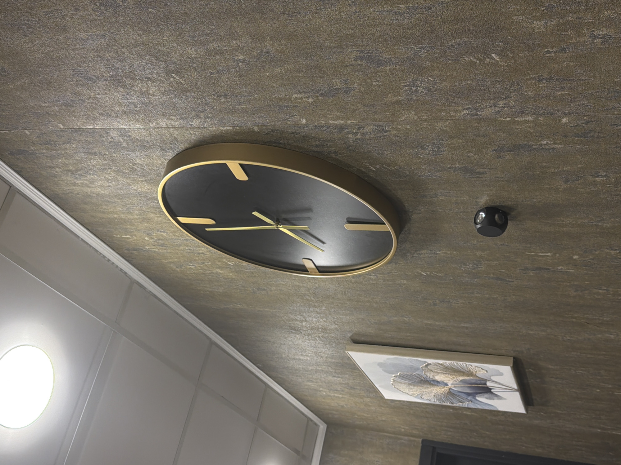 The image depicts a room interior in Podgorica, Montenegro. The ceiling has a textured brown surface with a linear pattern. Mounted on the ceiling is a black and gold circular clock with minimalist hour markers and golden hands, along with a small black device that appears to be a security camera or sensor. The lower right corner features a framed painting or artwork with an image of stylized leaves in shades of white, blue, and gold. The lower portion of the painting is partially obscured by a dark rectangle. A portion of a white wall with paneling is visible on the left side, illuminated by a round light fixture.