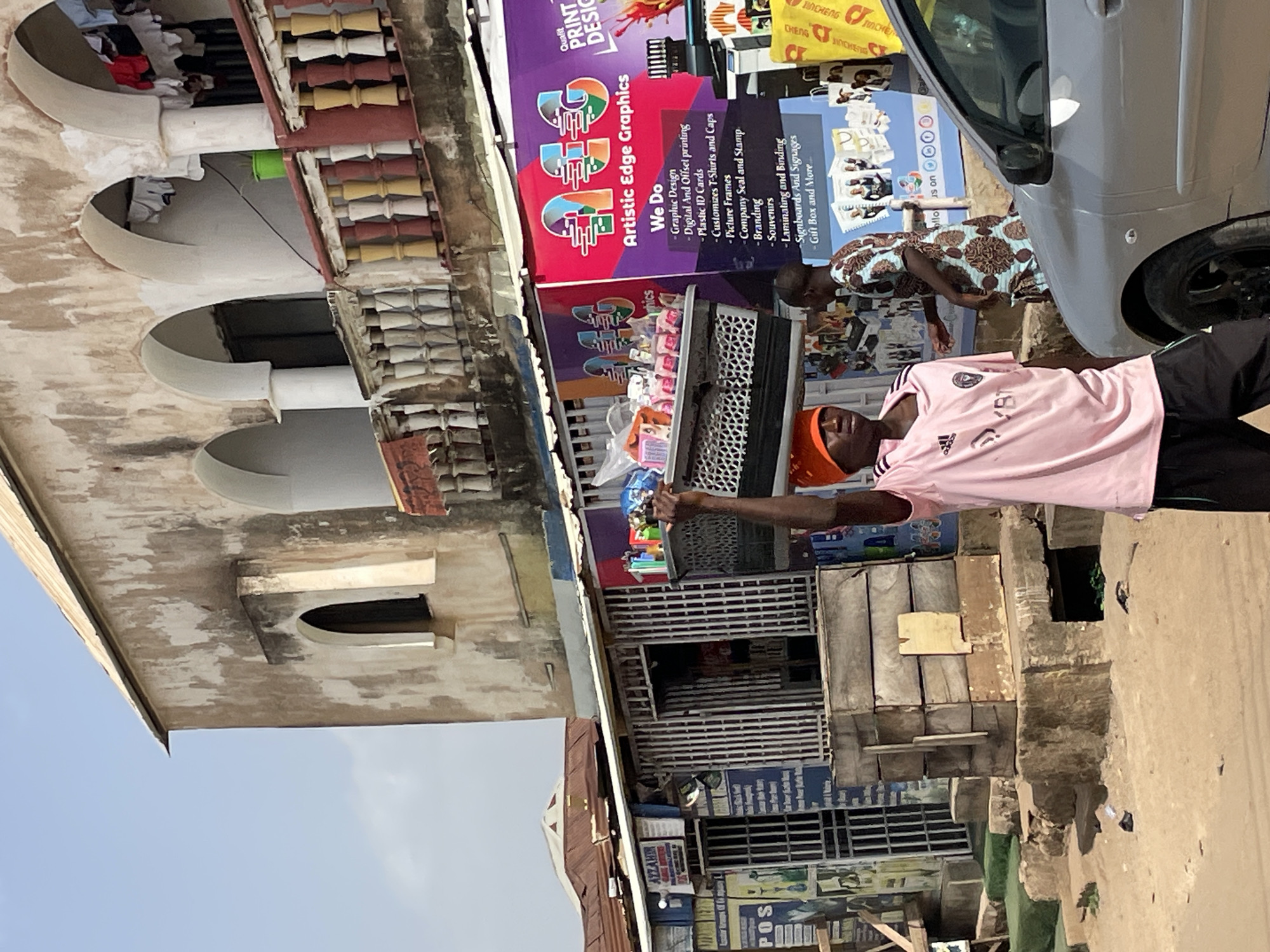 An outdoor scene in Jos, Nigeria, captures a bustling street with a mix of commercial activity and residential architecture under a clear daytime sky. In the foreground, a man in a pink Adidas t-shirt, dark pants, and an orange cap stands, looking towards the viewer and holding something in his extended right hand. The text "VBT" is visible on his shirt. To his right, partially obscured by a silver car, another person in a patterned blue and brown garment stands.
The street is unpaved and dusty, with various small, makeshift structures and what appears to be a metal grate or stall on the left. A silver car is parked near the right side of the frame. In the background, a multi-story building features distinctive arched window or balcony openings with elaborate balustrades, suggesting older architecture. Below this building, a vibrant purple and pink banner prominently displays "Artistic Edge Graphics," advertising services such as "Graphic Design," "Digital And Offset printing," "Plastic ID Cards," "Customizes T-Shirts and Caps," "Picture Frames," "Company Seal and Stamp," "Branding," "Souvenirs," "Laminating and Binding," "Signboards And Signages," and "Gift Box and More." Other signs with "JINCHENG" are visible, suggesting another business or product. The word "POS" is also visible on a small sign near the bottom right. The overall atmosphere is that of a lively urban commercial area.