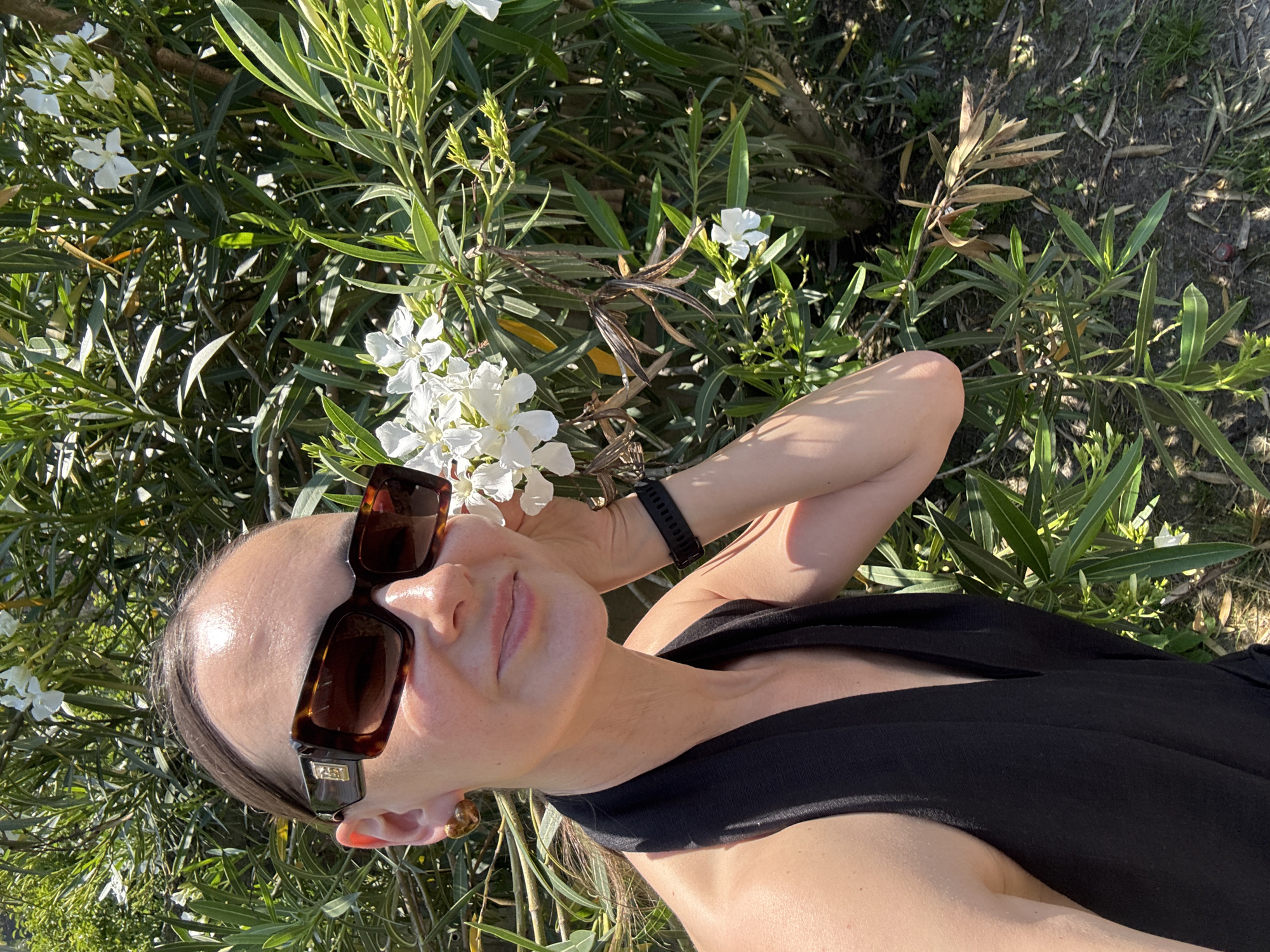 A woman with brown hair pulled back and wearing tortoiseshell sunglasses and a black halter top poses in front of a lush green bush with white flowers. She is wearing a gold earring and a black watch. The woman is smiling and her right hand is gently touching the white flowers. The scene appears to be outdoors on a sunny day. The image was taken in Sochi, Russia.