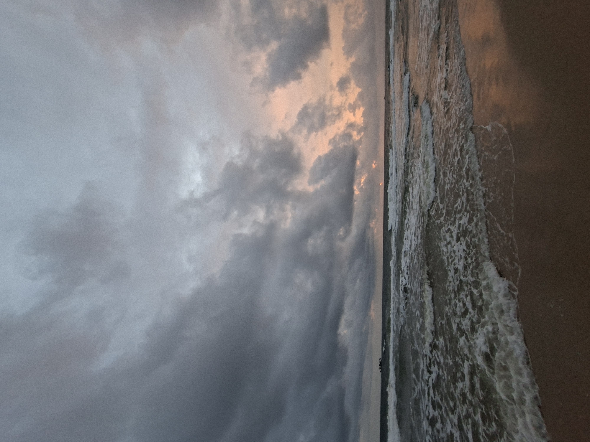 The image depicts a serene beach scene at dusk on Phú Quốc Island, Vietnam. The sky is filled with dramatic, dark gray clouds that are beginning to break, revealing a soft, peachy glow from the setting sun. The ocean is a deep, turbulent blue, with waves crashing onto the sandy shore. The sand itself is a warm, inviting brown, glistening with the receding tide. There are no people or man-made objects visible in the frame, emphasizing the natural beauty of the location. The overall mood is one of tranquil solitude, with the raw power of nature on full display.