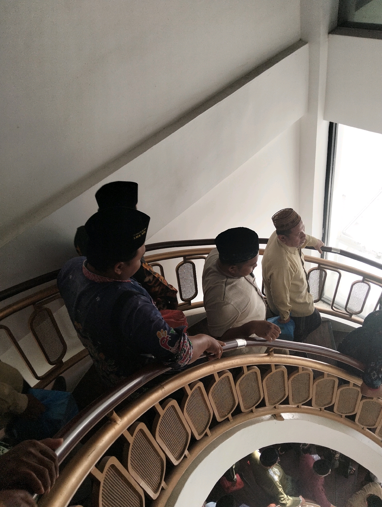 The image shows a spiral staircase in what appears to be a building in Paciran, Indonesia. The staircase has a golden railing with intricate lattice work. Four men are visible on the staircase, all wearing traditional Islamic attire, including peci hats. Two men are in the foreground, one wearing a dark blue shirt and the other a light-colored shirt. Two more men are further up the stairs, one wearing a light yellow shirt and the other a dark peci hat. Natural light streams in from a large window on the right side of the frame. Some people are also visible at the bottom of the staircase.