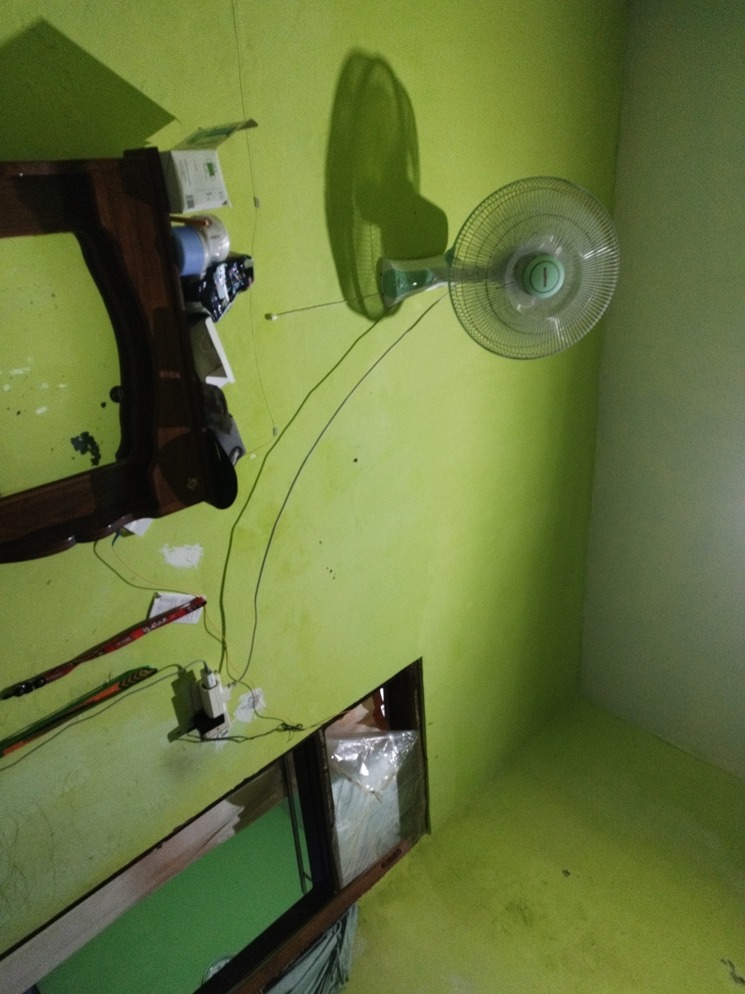 The image depicts an interior scene, likely a room, with walls painted a bright green. The orientation of the image suggests it was taken at an angle. Dominating the right side of the frame is a wall-mounted electric fan with a transparent protective grill. The fan is green and white. To the left, a wooden shelf unit with dark wood and some white elements is visible. On the shelf, there are several items, including what appears to be a roll of blue material, some white boxes or packaging, and dark objects. Below the shelf, on the wall, there are two colorful woven straps or belts. A white electrical outlet with a plug and wires attached is mounted on the wall below these straps. A dark, irregularly shaped opening or hole is present in the wall towards the bottom right of the image, revealing a glimpse of something covered in plastic sheeting within. The lighting in the room casts shadows, notably a large, distinct shadow of the fan on the wall behind it, and other shadows from the shelf unit and objects on it. The overall impression is of a simple, possibly residential, interior space. There are no people or animals visible in the image. The location is identified as Marengan Daya, Indonesia.