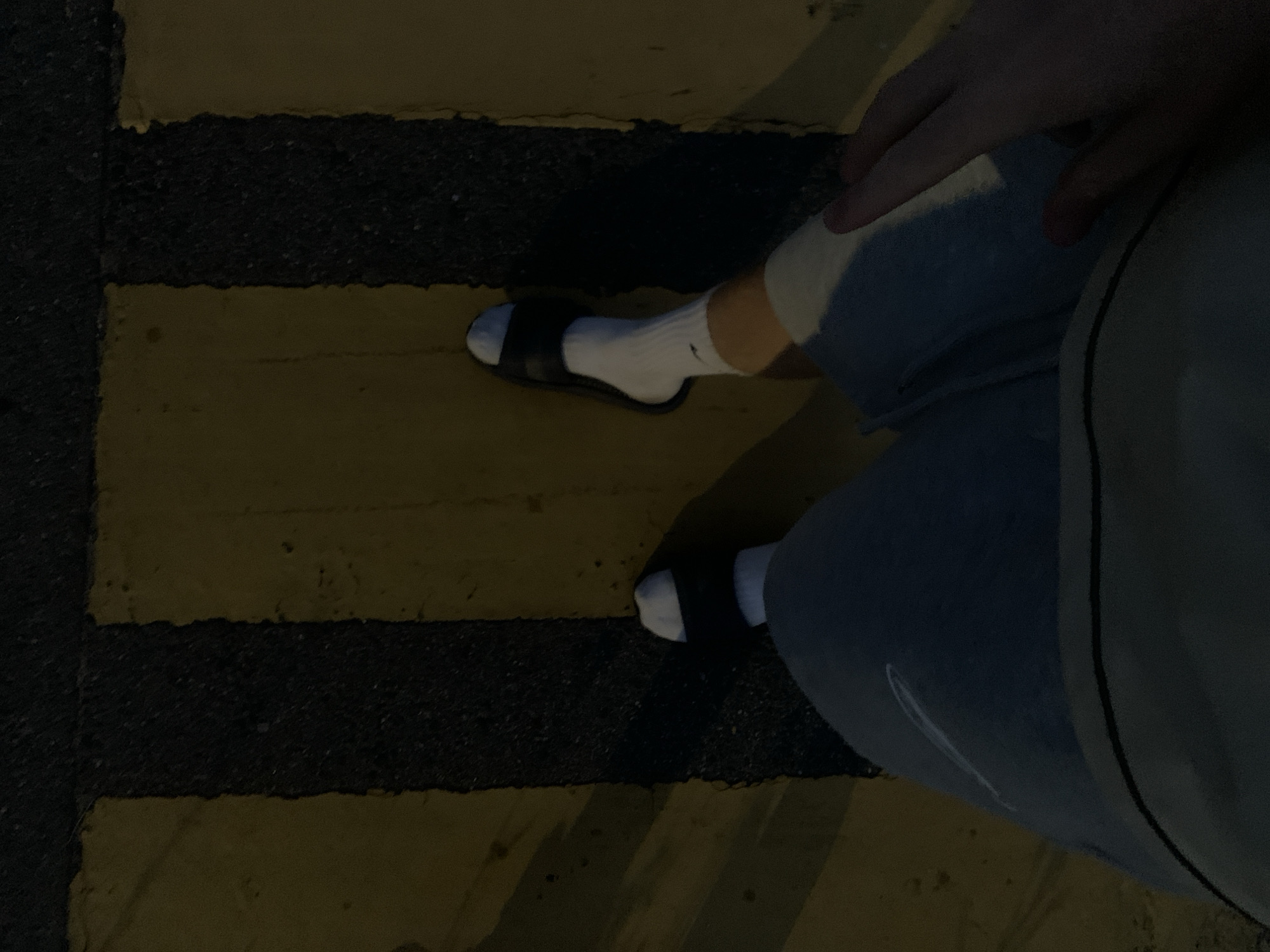 The image shows a first-person perspective of a person standing on a road with yellow and black striped markings, possibly a crosswalk or road delineation. The person is wearing light blue shorts, white socks, and black slide sandals. Their right hand is resting on their thigh. The lighting suggests it is nighttime or a dimly lit area. The provided crop hints do not offer any additional information about the specific location in Minsk, Belarus.