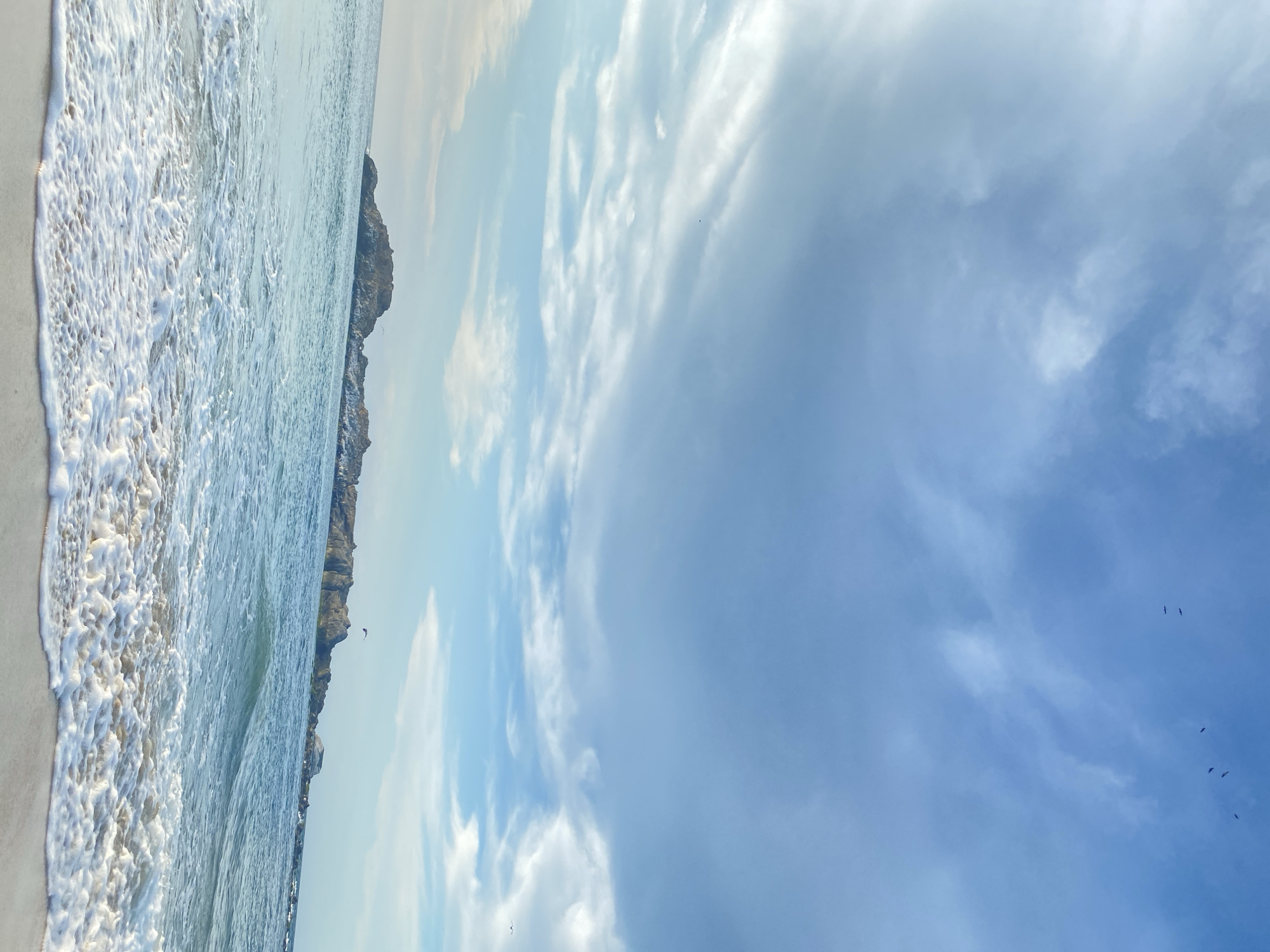 The image depicts a coastal scene, possibly near Altagracia, Venezuela, though this is based on contextual information and not explicit visual cues in the image. The left side of the frame features a beach with sand and frothy white waves crashing onto the shore. Adjacent to the water, a rocky cliff stretches into the distance. The right side of the image is dominated by a sky filled with light blue and white clouds. Several birds are visible as small, dark specks against the sky. The image orientation is rotated 90 degrees counter-clockwise.