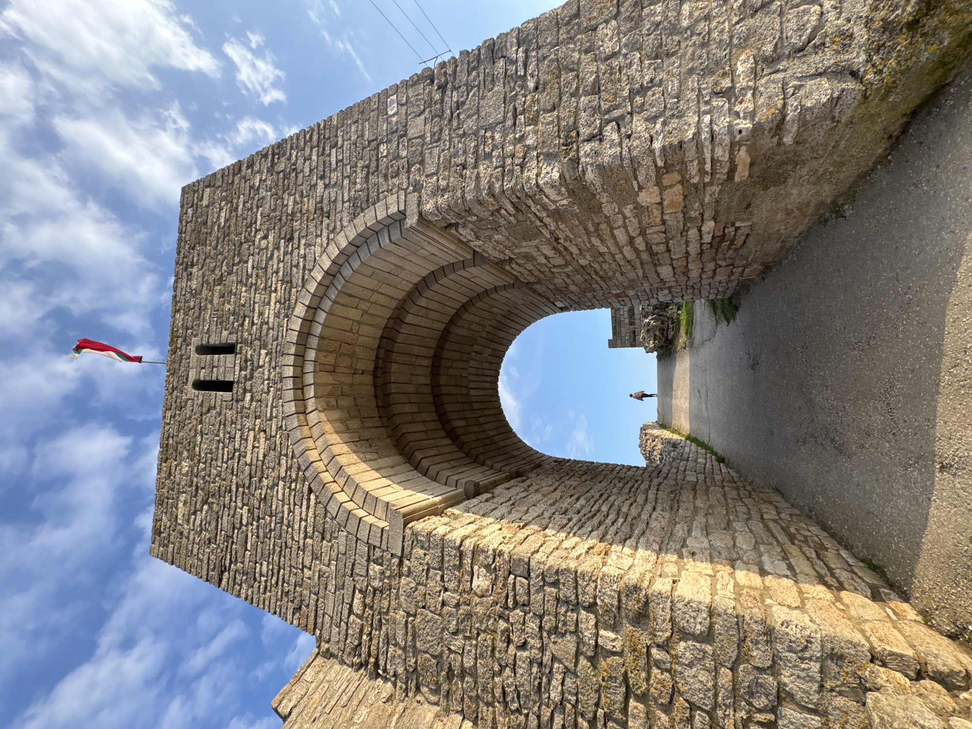 A low-angle shot depicts a stone archway. The arch is constructed of light-colored stones, forming a semi-circular opening. A red and white flag is affixed to the side of the arch. Through the archway, a narrow pathway is visible, leading to a distant figure standing on the path. The sky is blue with scattered clouds. The scene is located in Balgarevo, Bulgaria.