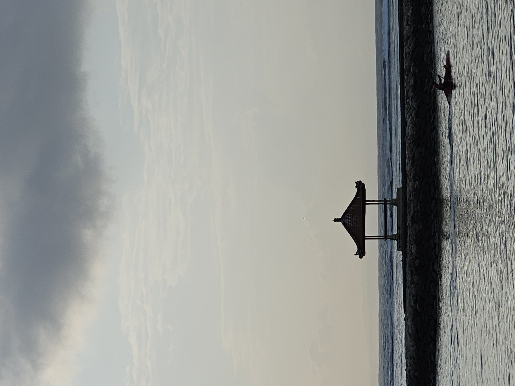 A wide shot captures a serene coastal scene in Denpasar, Indonesia. The image is dominated by a hazy sky with large, dark clouds obscuring the upper portion, giving way to a lighter, overcast sky with thin, streaky clouds. A small, ornate gazebo with a dark, pointed roof and open sides stands on a rocky pier that extends into the ocean. The pier is constructed from dark, irregular stones. Gentle waves ripple across the dark water in the foreground and background, indicating a calm sea. In the middle ground, a lone individual is kayaking on the water, facing away from the viewer, with an oar in their hands. The overall atmosphere is tranquil, with soft lighting due to the overcast conditions.