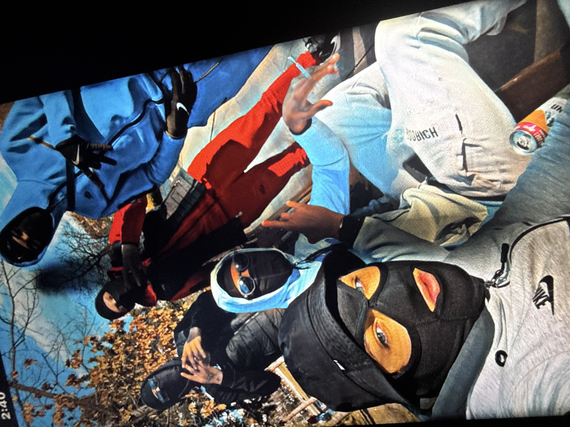 The image shows a group of young men wearing ski masks and hoodies, posing for a photo outdoors. One person in the foreground wears a black bucket hat and a black ski mask that covers their entire face except for their eyes and mouth. They are also wearing a grey Nike hoodie. To their left, another person in a black ski mask and black hoodie holds up a smartphone, possibly taking the picture. Behind them, a person in a red jacket and black ski mask stands next to someone in a bright blue tracksuit and black gloves with white Nike logos. Another individual in a light blue hoodie and sunglasses with a black ski mask is visible in the middle of the frame. The background appears to be an outdoor setting with trees and what looks like a wooden bench. The overall atmosphere suggests a casual and informal gathering. The timestamp in the upper left corner indicates 2:40. The specific location in Sofia, Bulgaria, cannot be determined from the visual information provided.