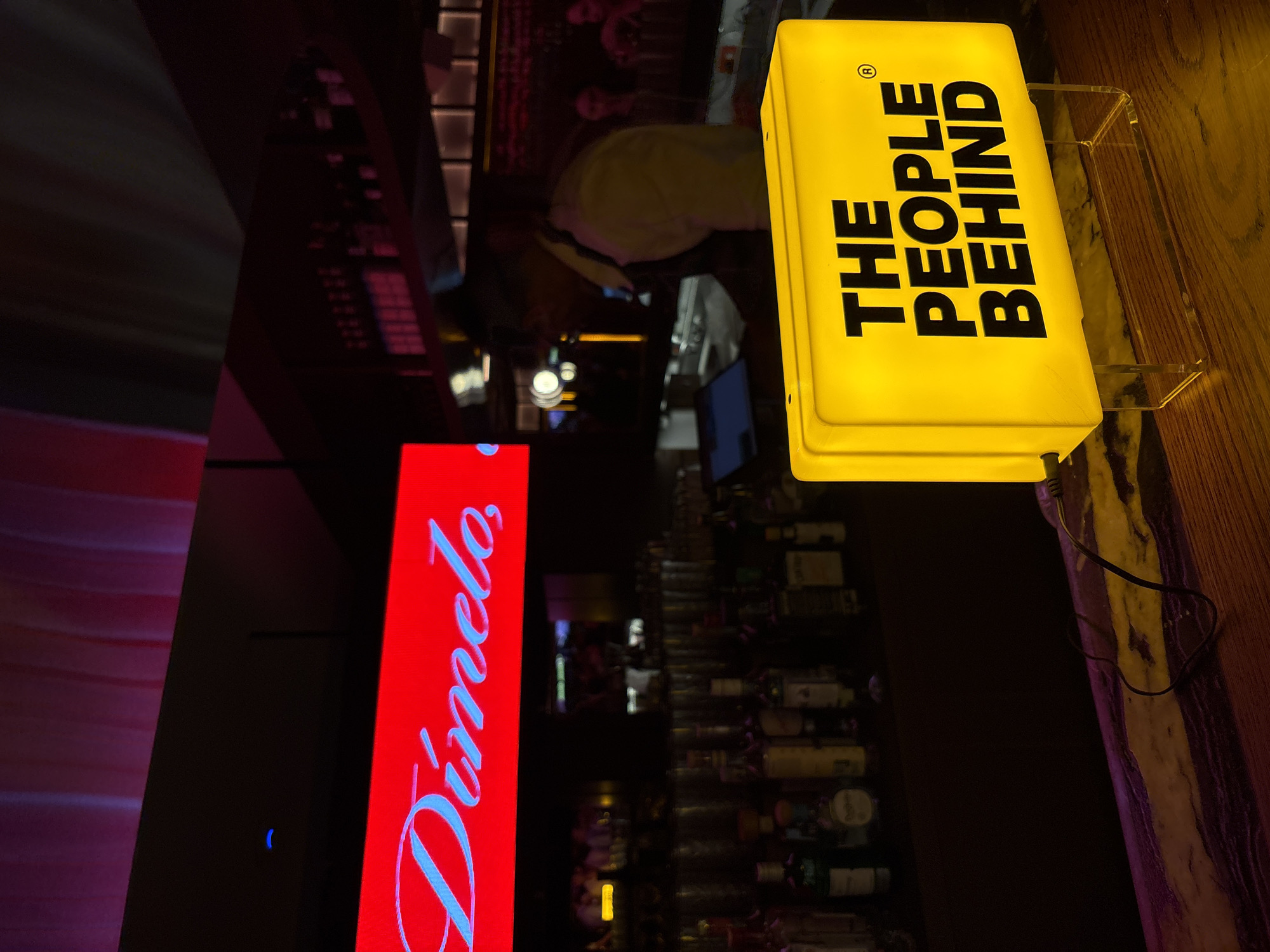 An indoor, low-light photograph, rotated 90 degrees clockwise, depicts a bar or similar establishment. The scene is dominated by two brightly illuminated signs and a person working behind a counter.
In the upper right quadrant of the image (when oriented correctly), a rectangular yellow illuminated sign is prominently displayed. It features black uppercase text that reads "THE PEOPLE BEHIND" stacked in two lines, with a registered trademark symbol (®) in the top right corner. The sign appears to be resting on a wooden counter or surface.
To the left of the yellow sign and extending into the middle left, a tall, vertical red illuminated sign is visible. This sign displays the word "Dínelo" in a stylized, light blue or white script font.
Behind the signs and across the background, a person, likely a bartender, is partially visible, wearing a light-colored top. They are situated behind what appears to be a bar counter, with numerous bottles arranged on shelves or racks in the background. The bottles are mostly obscured by the dim lighting but suggest a well-stocked bar. The overall ambiance is dark, with the illuminated signs providing the primary light sources, hinting at a nighttime setting or a venue with atmospheric lighting.