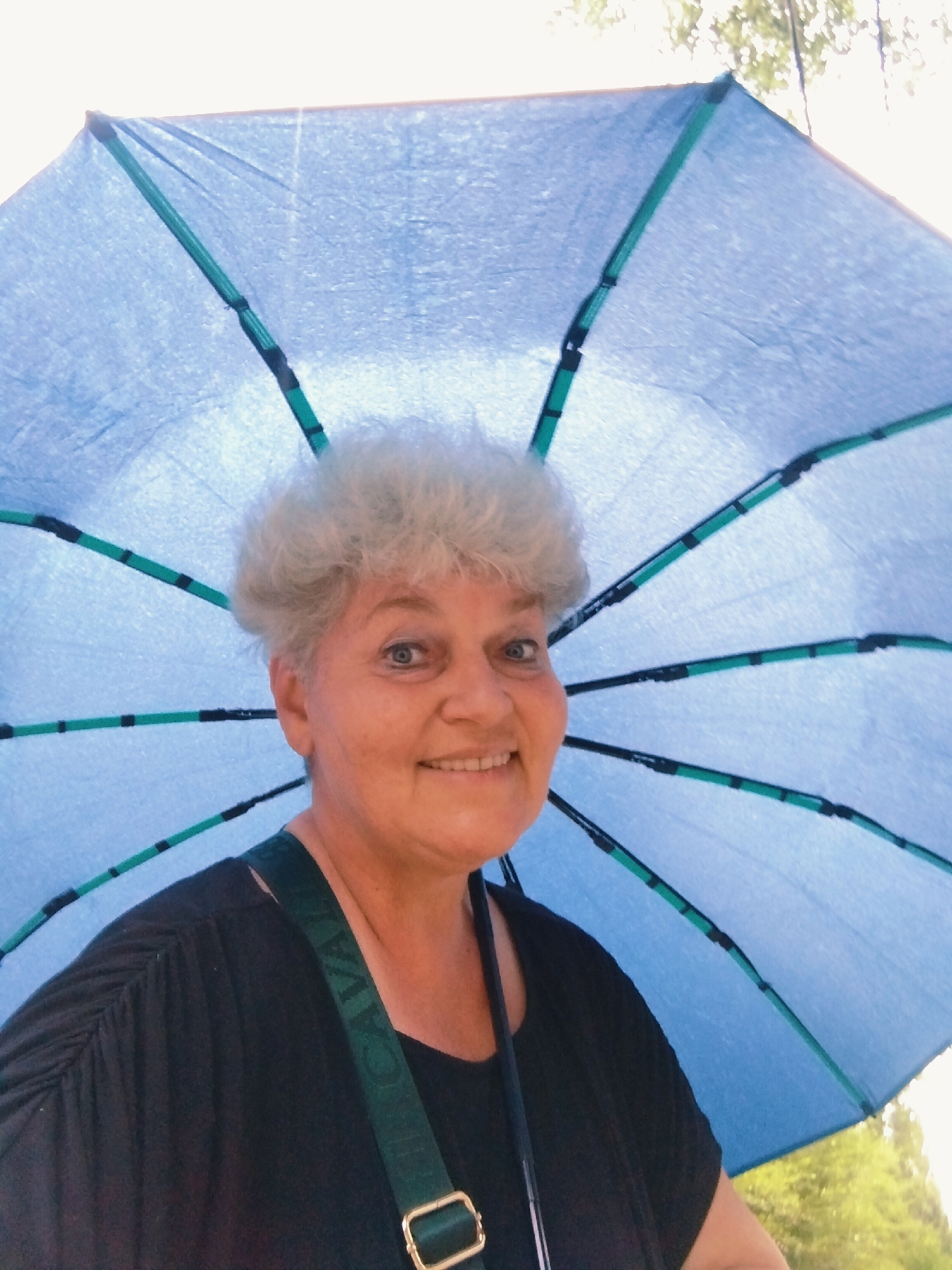 An outdoor, waist-up, eye-level, medium shot captures an older woman with light-colored, short, curly hair, holding a bright blue umbrella behind her. She wears a black shirt and a green strap with gold hardware crosses her chest. The woman is smiling at the camera, and her head is tilted slightly to her left. The umbrella, with its green ribs, is open and casts a slight shadow over her face. The background is blurred, with hints of green foliage and sunlight filtering through trees, suggesting a park or natural setting. The overall lighting is bright, indicating daylight.