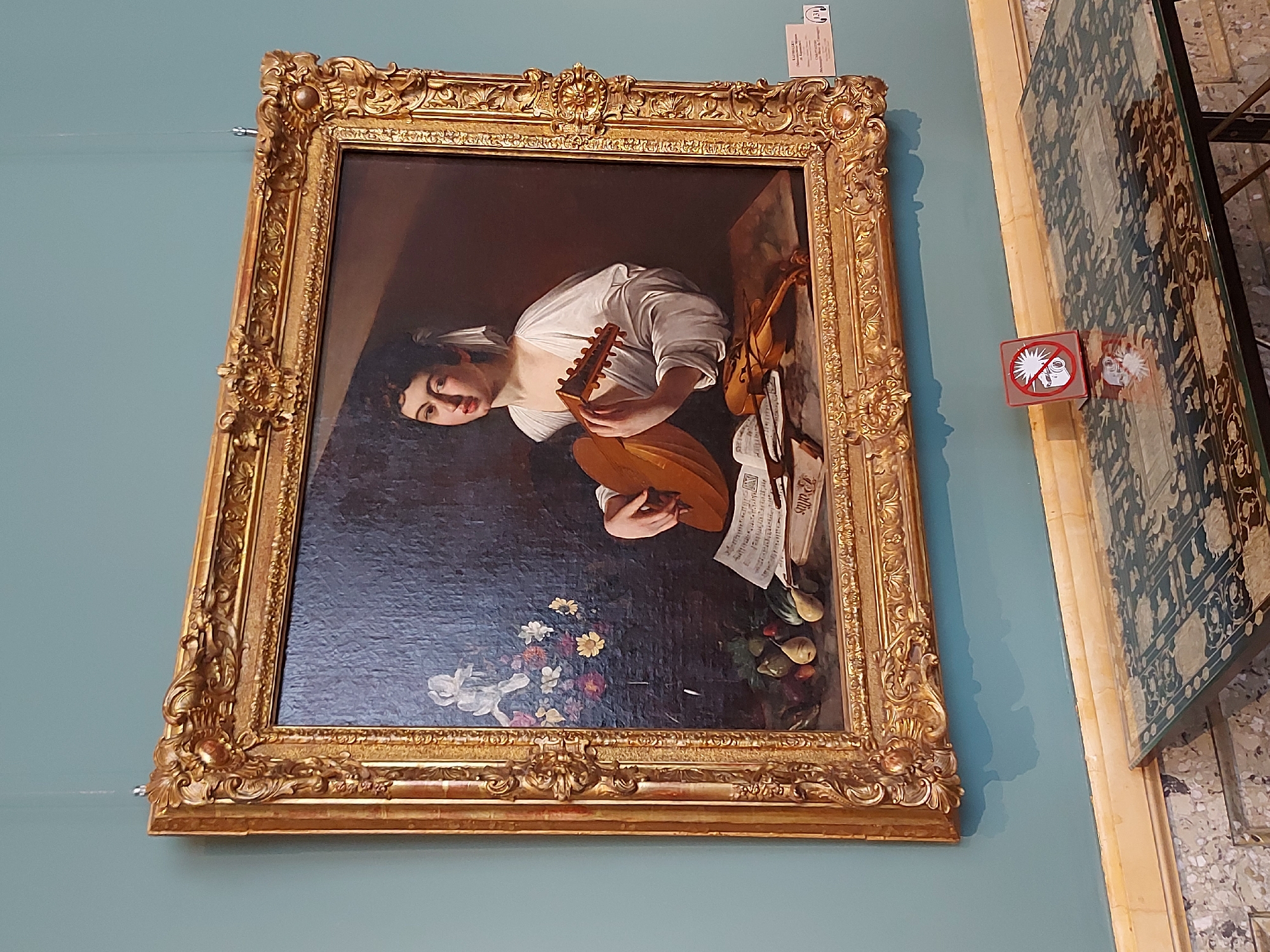 The image depicts a framed painting of a woman playing a lute. The painting is in an ornate gold frame. The woman in the painting is wearing a white dress. In the background, there is a dark space with some flowers and fruit at the bottom. To the right of the painting, there is a "no flash photography" sign and a decorated table. The painting is displayed on a blue wall. The city and specific location are unknown.