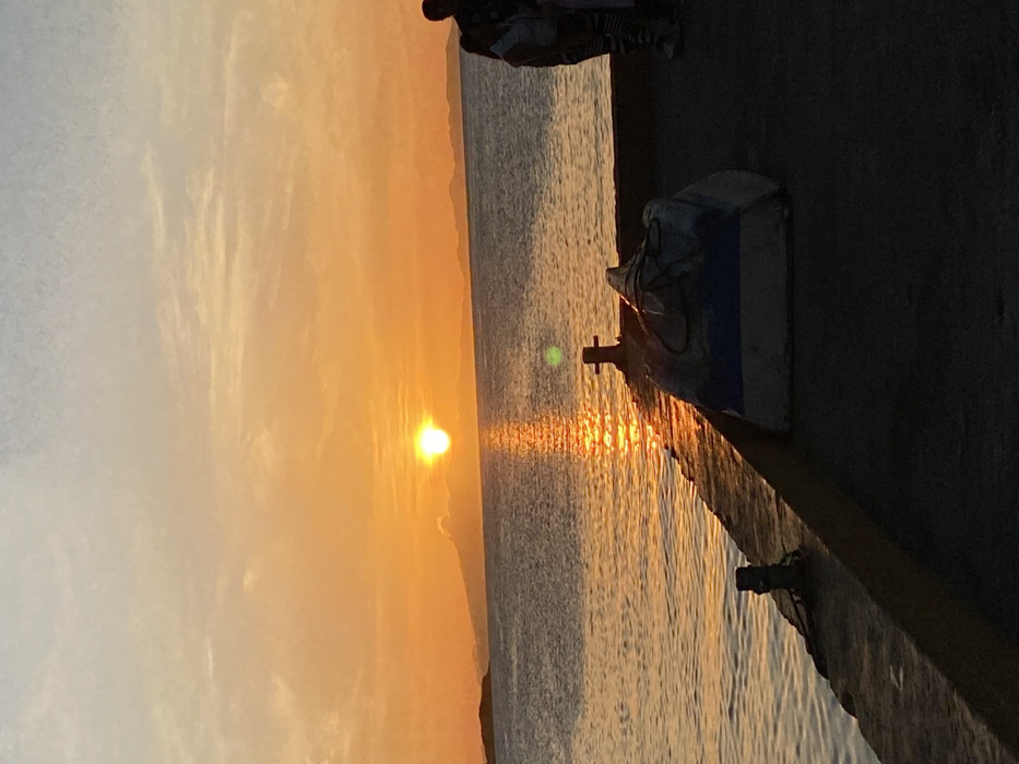 A serene landscape depicts a vibrant sunset illuminating the sea. The sun, low on the horizon, casts a golden glow on the water and sky. A stone pier stretches into the sea, with a small boat docked alongside. The pier is weathered, and the boat appears old and slightly dilapidated. The sky is filled with orange and yellow hues, fading into softer shades near the horizon.
