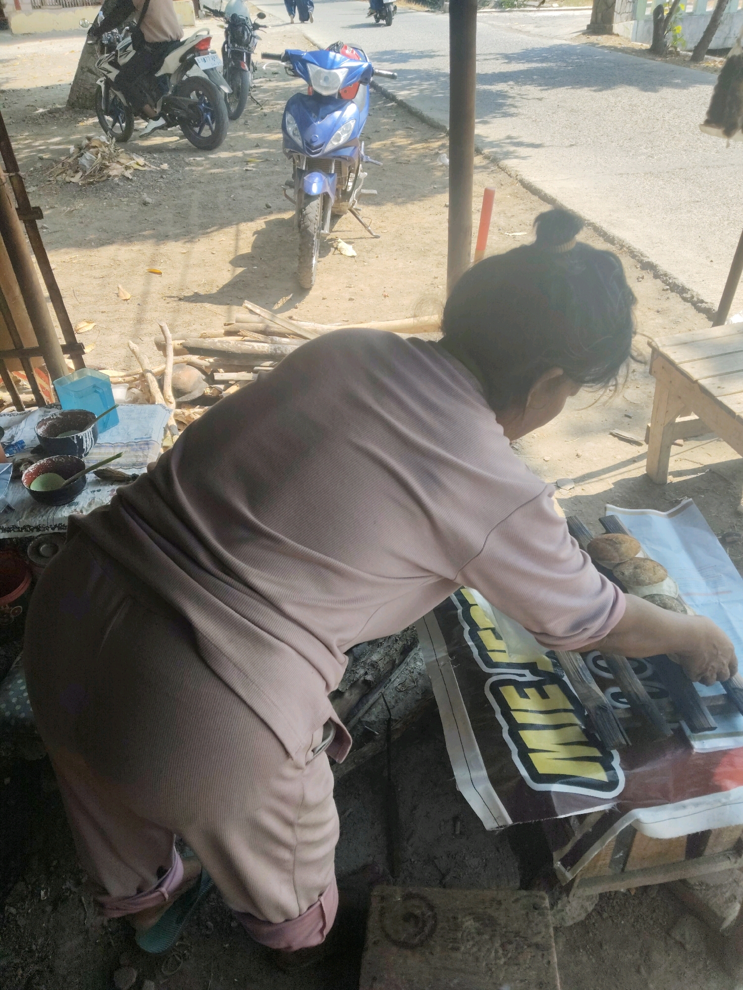 A medium shot shows a person in light pink clothing preparing food outdoors in Subang, Indonesia. The person, facing away from the camera and hunched over a makeshift table, is using a brush to apply something to what appear to be four round food items arranged on a wooden plank. These items, possibly bread or cakes, are resting on a white and brown tarp that has a yellow and black logo partially visible. The table itself is covered by the tarp and is situated on the ground, which is dirt and debris. Several bowls and utensils are on a blue cloth to the left of the frame. Behind the person, a blue motorcycle is parked, and further in the background, another motorcycle with a rider is visible on a paved road. The scene is illuminated by sunlight, casting shadows on the ground. The background includes trees, a fence, and what appears to be a building or structure.