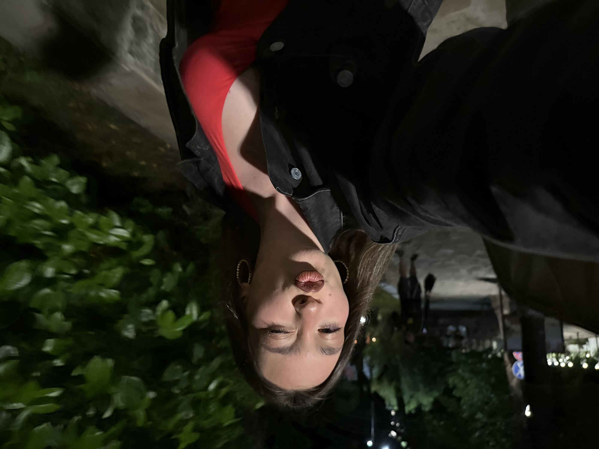 This nighttime outdoor selfie depicts a young woman in Sozopol, Bulgaria, against a dimly lit background of trees and urban elements. The woman, positioned centrally, faces the camera with her left arm extended, holding the device. She has long, dark brown hair and is shown winking her right eye while puckering her lips with dark brown lipstick. She wears gold-toned hoop earrings and a black denim jacket unbuttoned over a vibrant red top with a V-neckline, revealing a portion of her chest.
In the left-to-mid background, a blurred male figure is visible walking away, wearing a dark top and shorts, carrying a dark object. Further back, numerous blurred yellow-white lights suggest distant streetlights or decorative illumination. A partially obscured circular sign, featuring blue and red segments, is situated behind the woman's left shoulder. To the right, dense green foliage occupies the mid-ground and extends upwards. A low, light-colored stone or concrete wall runs horizontally along the bottom right edge of the frame. The overall scene is characterized by low light conditions typical of an evening or night setting.