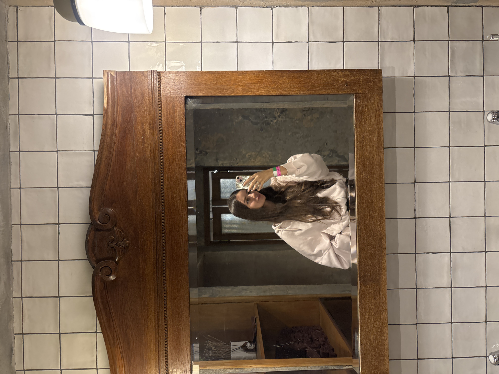The image shows a mirror selfie of a woman in Moscow, Russia. The mirror is large with a wooden frame, featuring decorative carvings on the left side. The background reflected in the mirror includes a window, shelves with various items, and what appears to be a concrete interior. The woman is holding a phone and wearing a light-colored jacket or robe. The bathroom or room where the mirror is located has white square tiles with dark grout on the walls. A light fixture is visible above the mirror.
