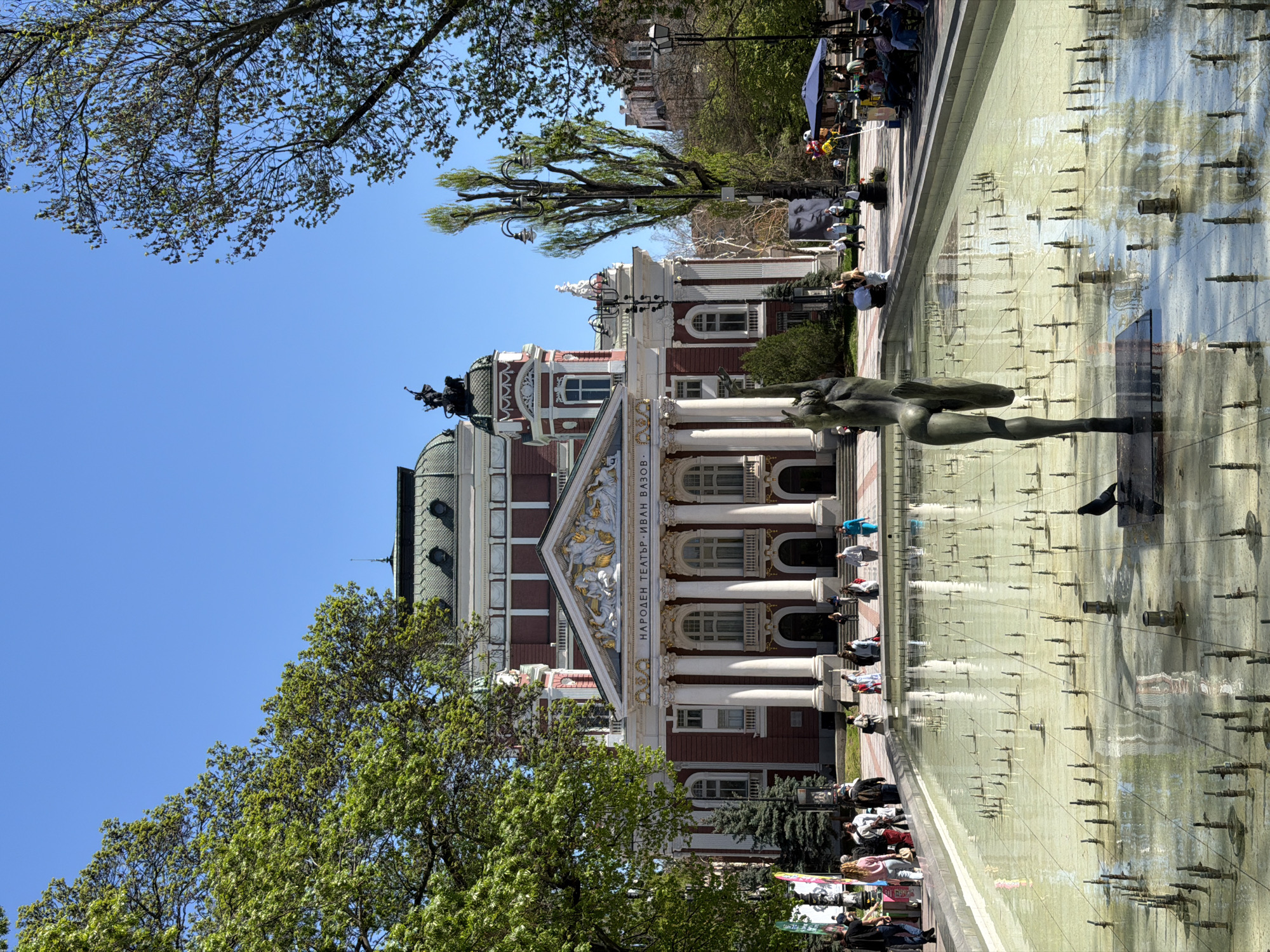 The image captures a sunny day in Sofia, Bulgaria, at the square in front of the Ivan Vazov National Theatre. A large rectangular fountain stretches across the foreground, featuring numerous small water jets. Adjacent to the fountain, the classical facade of the theatre is visible, with its white columns and detailed architectural ornamentation. People are gathered on the steps of the theatre and alongside the fountain, some sitting and others standing. In the foreground is a bronze statue. Trees with green foliage frame the scene, providing a natural border. The sky is clear and blue.