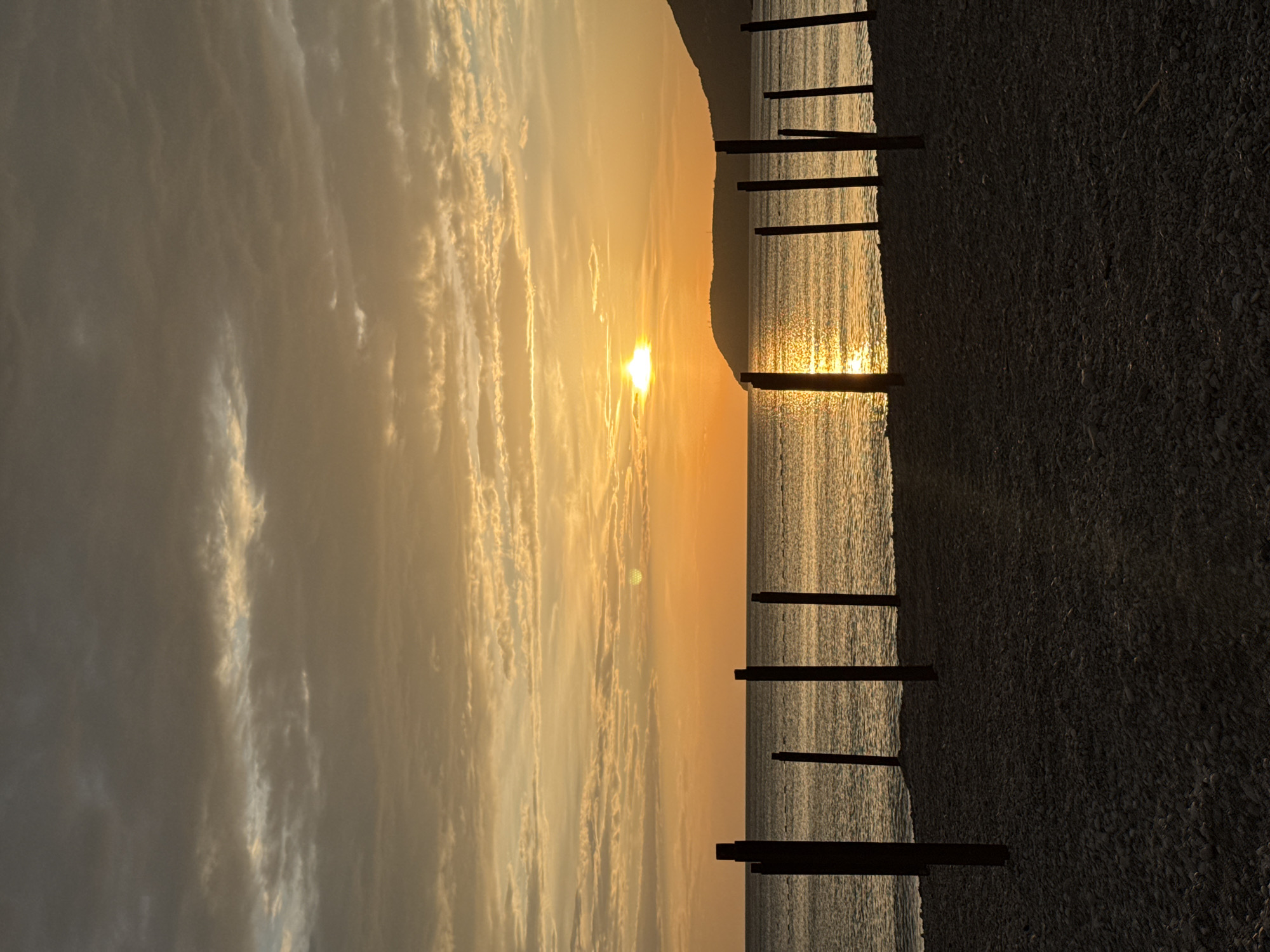 The image captures a sunset over the Adriatic Sea in Šušanj, Montenegro. The sun is partially obscured by a mountain range in the distance, casting a golden glow on the water and sky. The sky is filled with streaks of clouds, ranging in color from gray to gold. Along the shoreline, a series of wooden posts stand partially submerged in the water. The beach itself is composed of small pebbles and is largely in shadow. No people are visible in the image. The dominant colors are gold, gray, and brown.