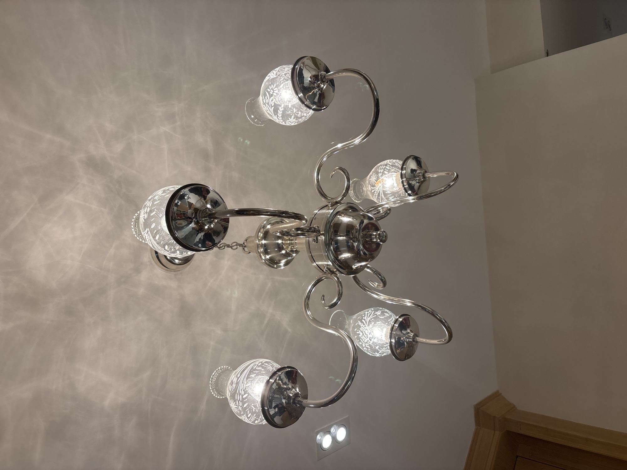 The image displays an interior room in Mexico City, Mexico, featuring a five-arm polished chrome chandelier centrally mounted on a white ceiling. Each arm supports an illuminated, upward-facing light fixture encased in a clear glass shade etched with a floral motif. Light from these fixtures projects distinct patterns onto the ceiling. Two illuminated recessed spotlights are present in the upper-left of the ceiling. The lower-left corner reveals a segment of a light-colored wooden architectural trim piece adjacent to a plain, light-toned wall. No individuals or direct interactions are depicted; the scene is illuminated by artificial lighting.