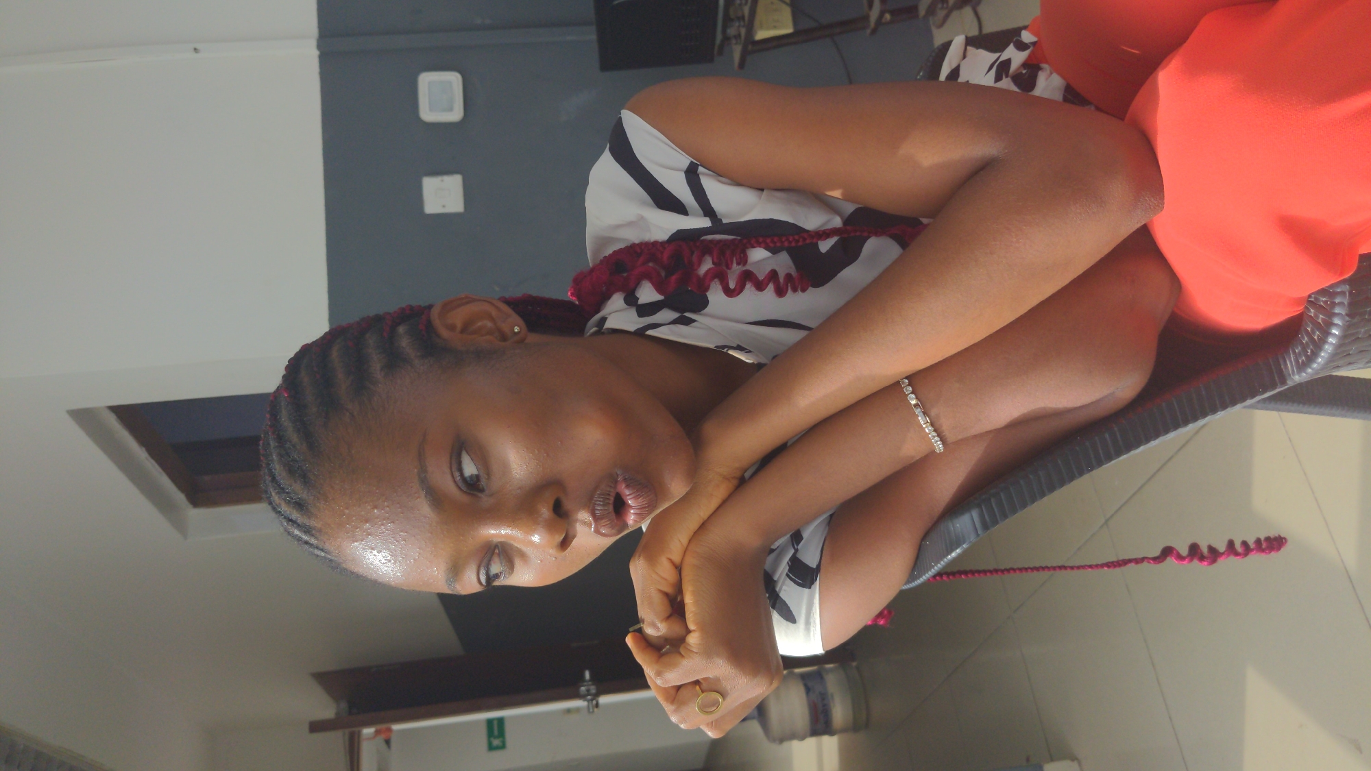 A young Black woman is pictured seated in an indoor setting, likely in Abuja, Nigeria. She is wearing a sleeveless top with a black and white abstract pattern and a vibrant red-orange skirt or dress. Her hair is styled in dark cornrows, intricately braided with some red/maroon curly extensions visible around her neck and hanging down to the floor. She has a silver-colored bracelet on her left wrist and a simple gold-toned ring on her right ring finger.
The woman is seated in a dark gray, possibly rattan or plastic, chair. She is leaning forward, with her arms crossed and her hands clasped together. Her head is tilted slightly, and her lips are parted in an expressive manner, suggesting she might be speaking, listening intently, or conveying a particular emotion like curiosity or contemplation. Her gaze is directed towards her left.
The background consists of a two-toned wall, with a light gray or white section on the left and a darker gray section on the right, featuring a light switch or electrical outlet. A square opening, possibly a window or a decorative architectural detail, is visible in the upper part of the white wall. On the floor behind her, a water dispenser or cooler can be seen. The overall lighting suggests it is daytime. There is no visible text in the image.