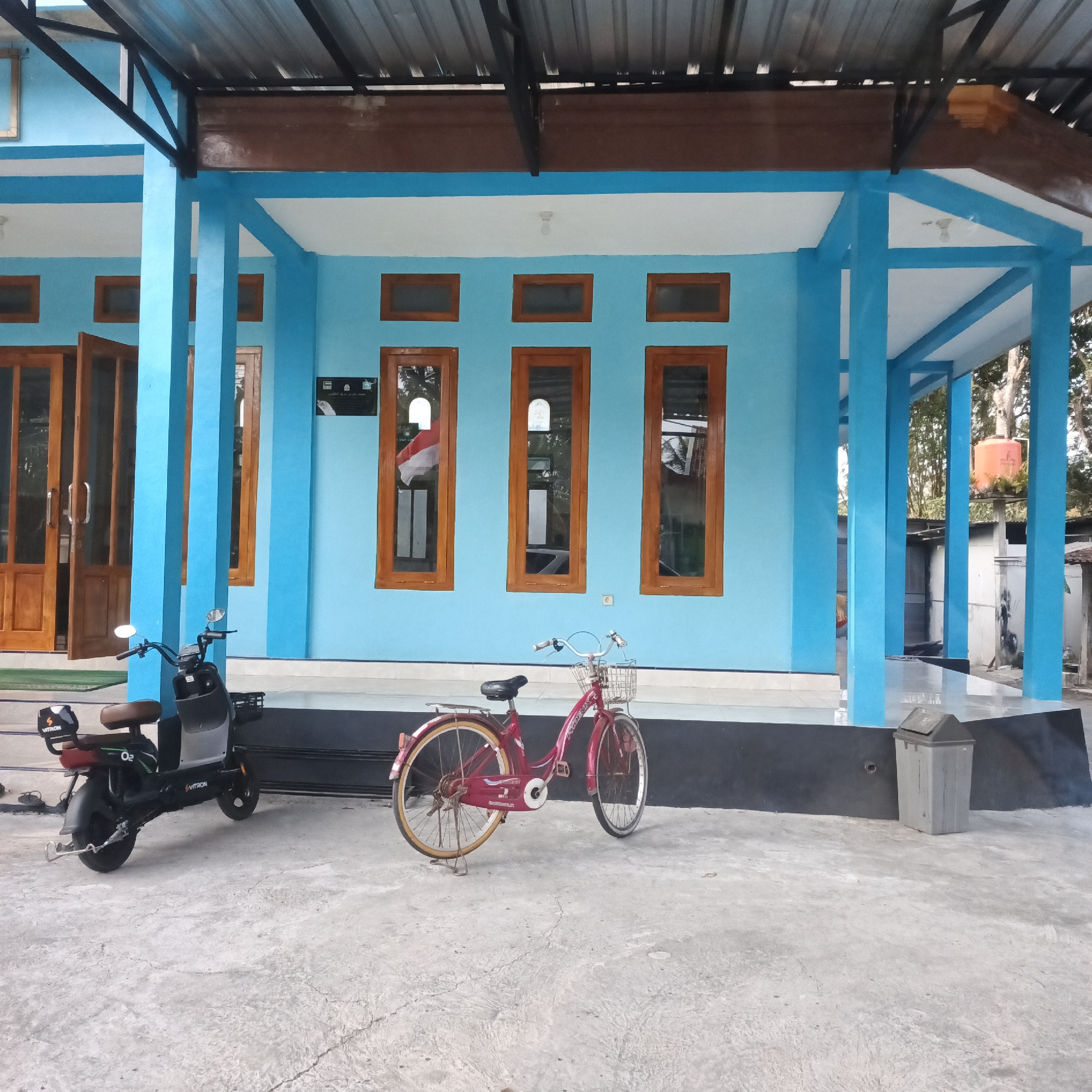 In Karangtalun, Indonesia, a bright blue building with a corrugated metal roof supported by blue columns is depicted. In front of the building, on a concrete ground, an electric scooter is parked on the left and a red bicycle with a basket is on the right. The building features several wooden-framed windows, some of which reflect a red and white flag, likely the Indonesian flag. The scene appears to be an outdoor area, possibly a school or community building, with trees visible in the background. A gray trash can is positioned near the front of the building. The overall impression is one of a simple, functional public space.