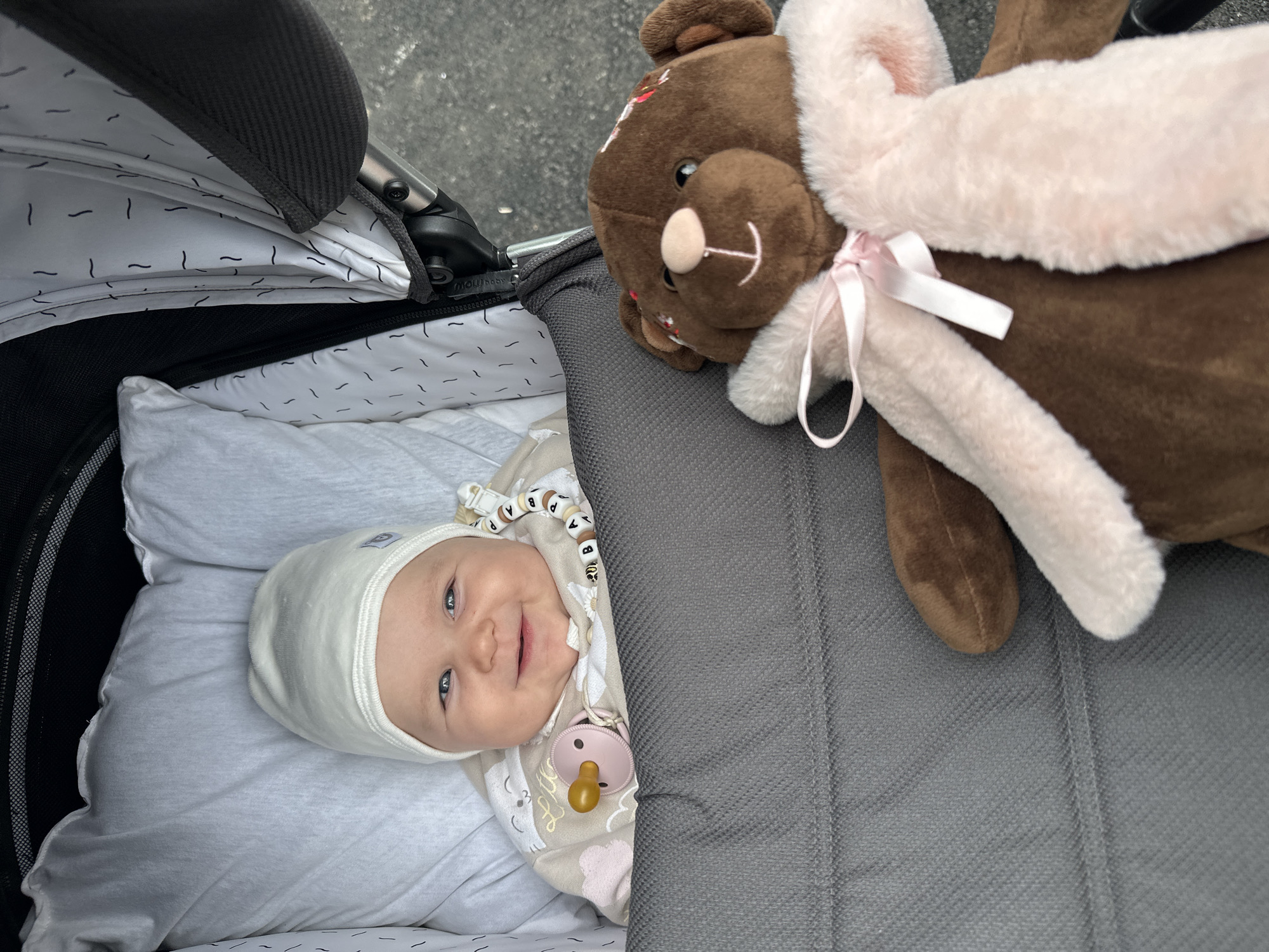 The image shows a baby in a stroller, next to a stuffed bear. The baby is wearing a white hat and a light-colored onesie with a pacifier attached. The stroller is gray with a patterned lining. The stuffed bear is brown with a pink scarf. The background appears to be an outdoor setting, possibly a street or sidewalk, with asphalt visible. The image does not contain any information indicating the location in the city of Chaikovsky, Russia.