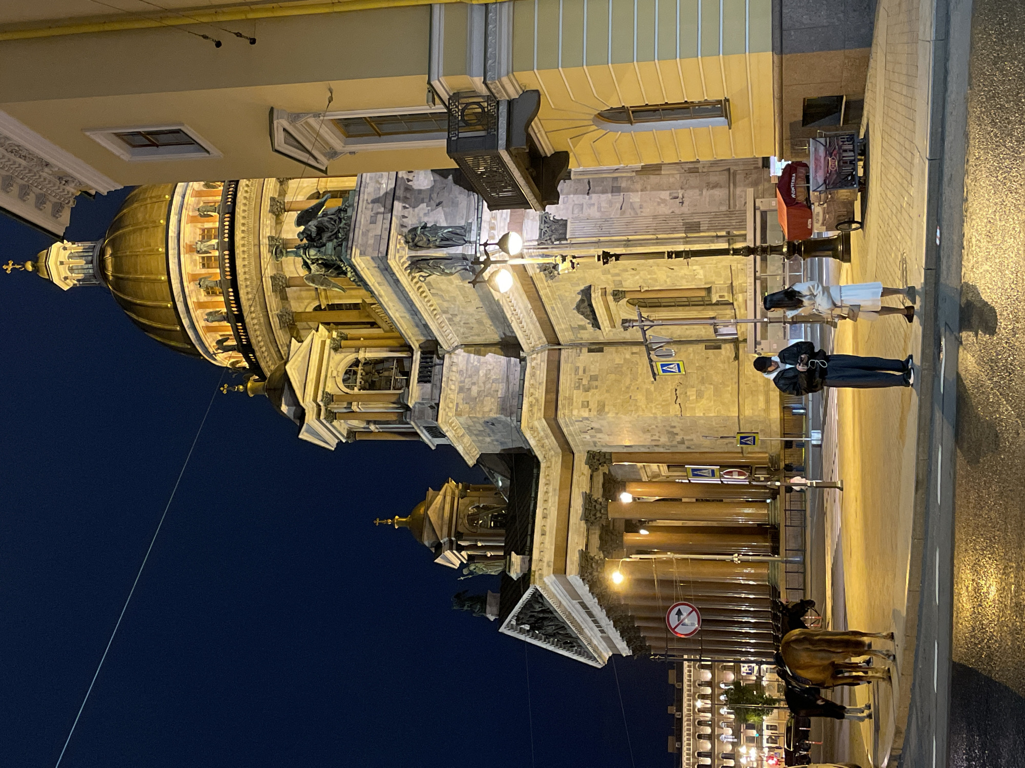The image shows a nighttime view of St. Isaac's Cathedral in St. Petersburg, Russia. The cathedral's golden dome dominates the scene against a dark blue sky. The building is brightly illuminated, highlighting its architectural details and statues. Two people are standing on the sidewalk, and a horse-drawn carriage is visible near the cathedral's columns. The street is wet, reflecting the ambient light. Street signs and lights are also visible.